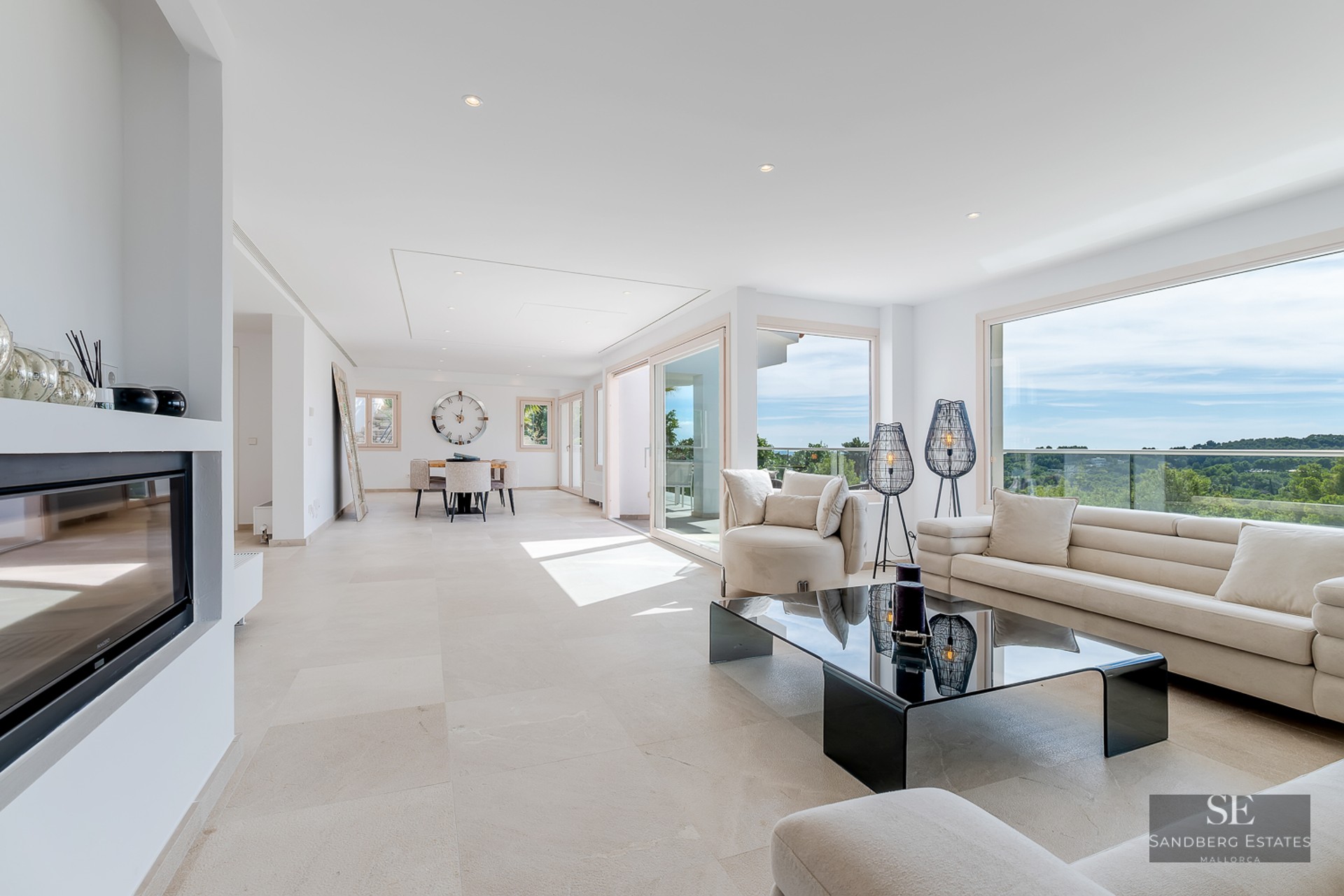 White living room with beige sofas, glass coffee table, fireplace, and large windows overlooking a green landscape.