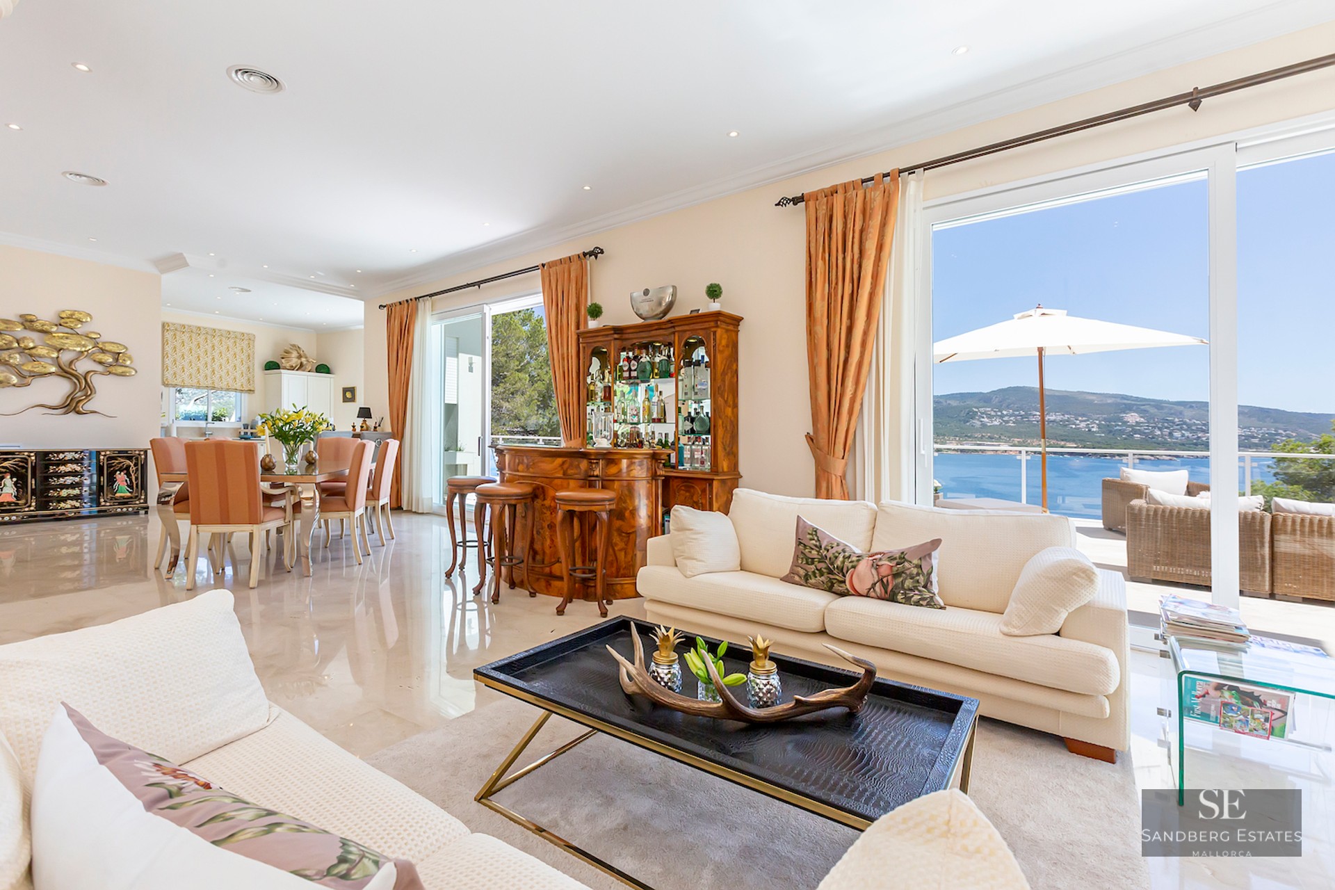 Spacious living room with cream sofas, classic wooden bar, dining area, and large glass doors overlooking the sea.