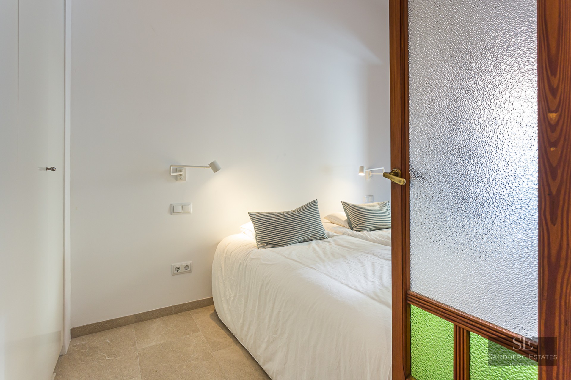 Modern bedroom view through a wooden door with textured glass, featuring a white bed and wall-mounted reading lamps.