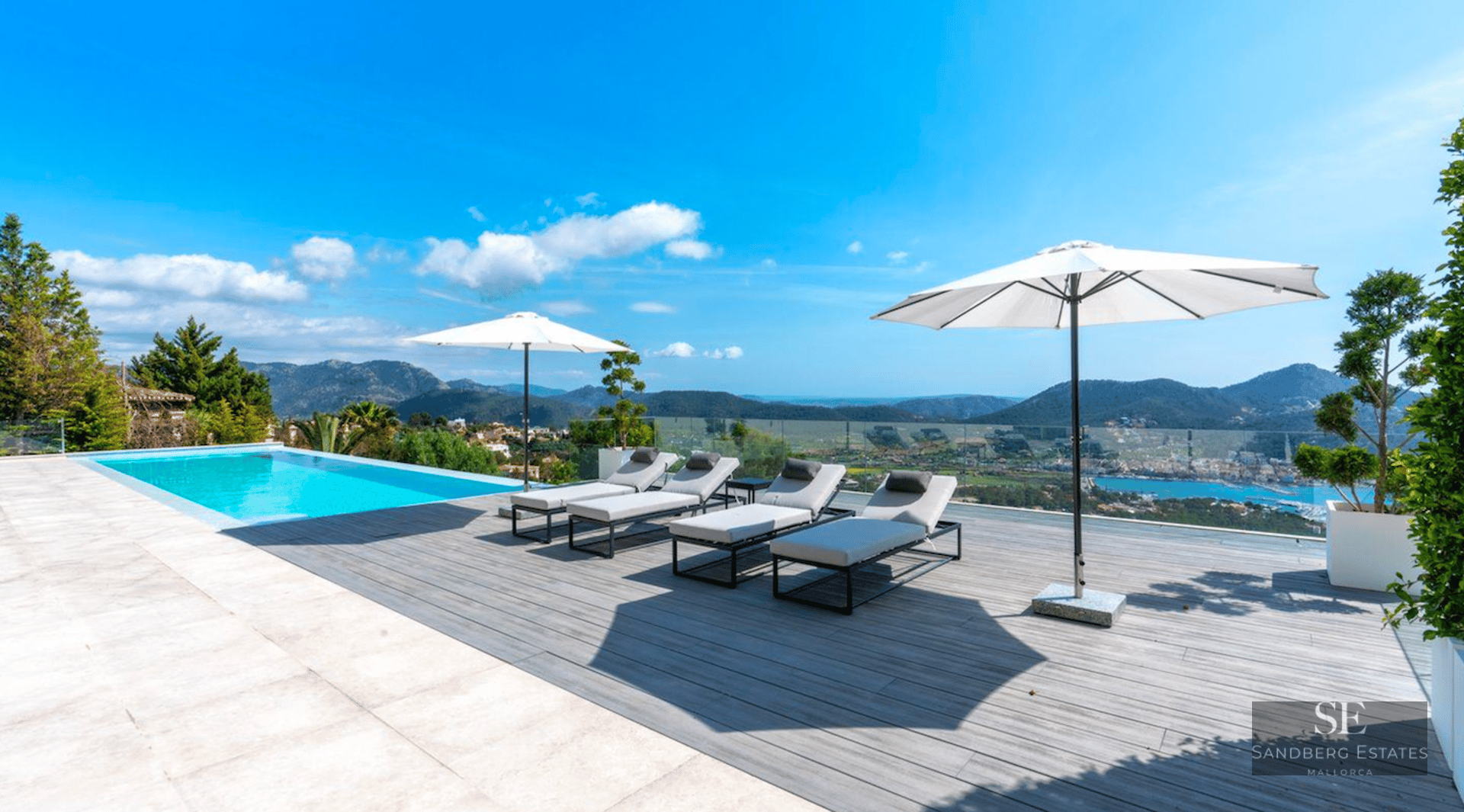 Infinity pool on a wooden deck with grey loungers, white umbrellas, and a vast view of mountains and the sea.