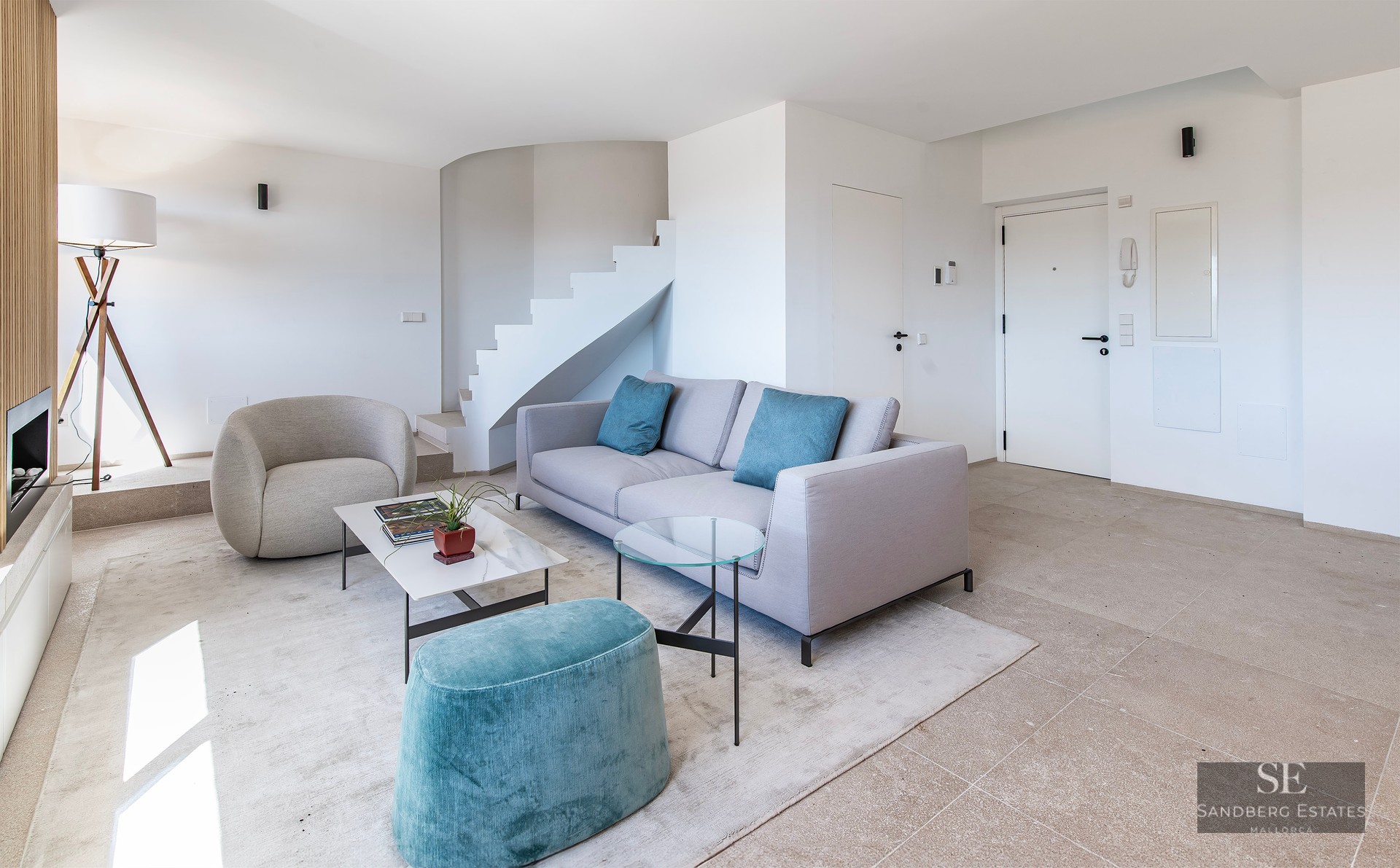 Bright minimalist living room with grey sofa, teal ottoman, white staircase, and marble coffee table.