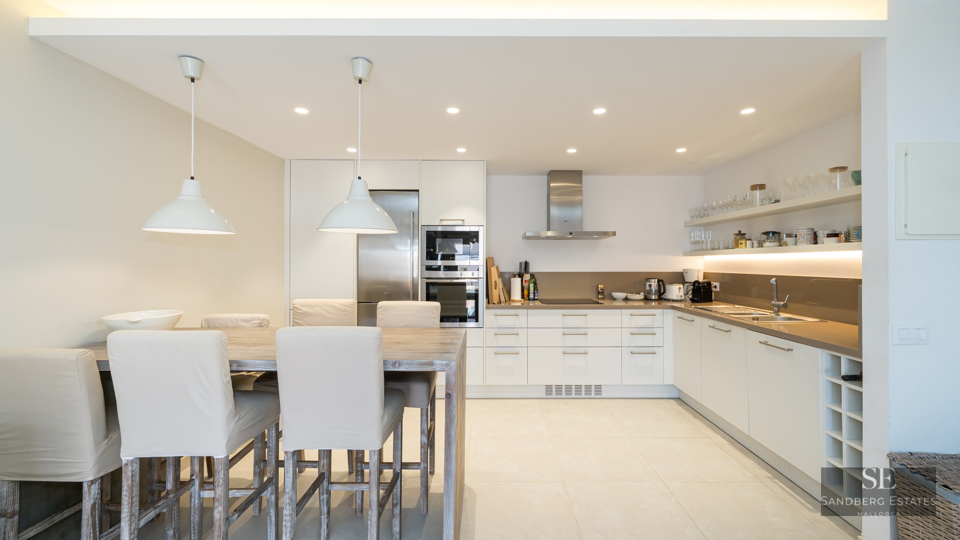 A bright modern kitchen featuring white cabinets, stainless steel appliances, and a large wooden dining table with cream chairs.