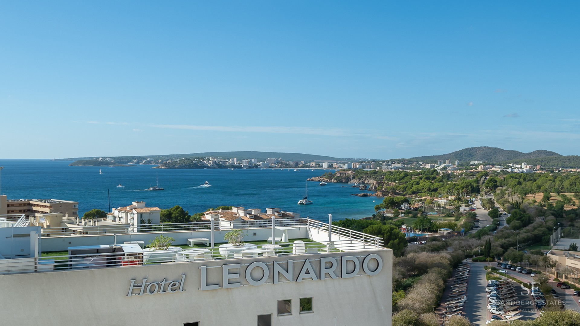 Panoramic view of a Mediterranean bay and coastline from a modern rooftop terrace with white railings and artificial turf.