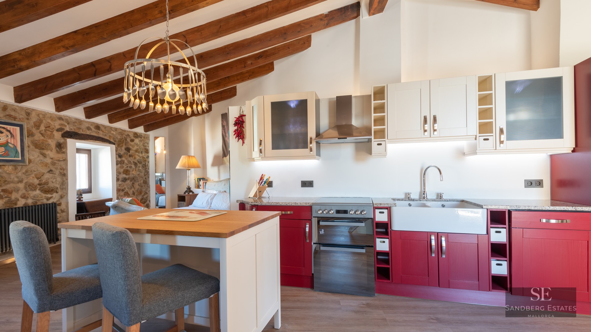 Rustic kitchen featuring red cabinets, stone walls, and exposed wooden ceiling beams with a unique spoon chandelier.