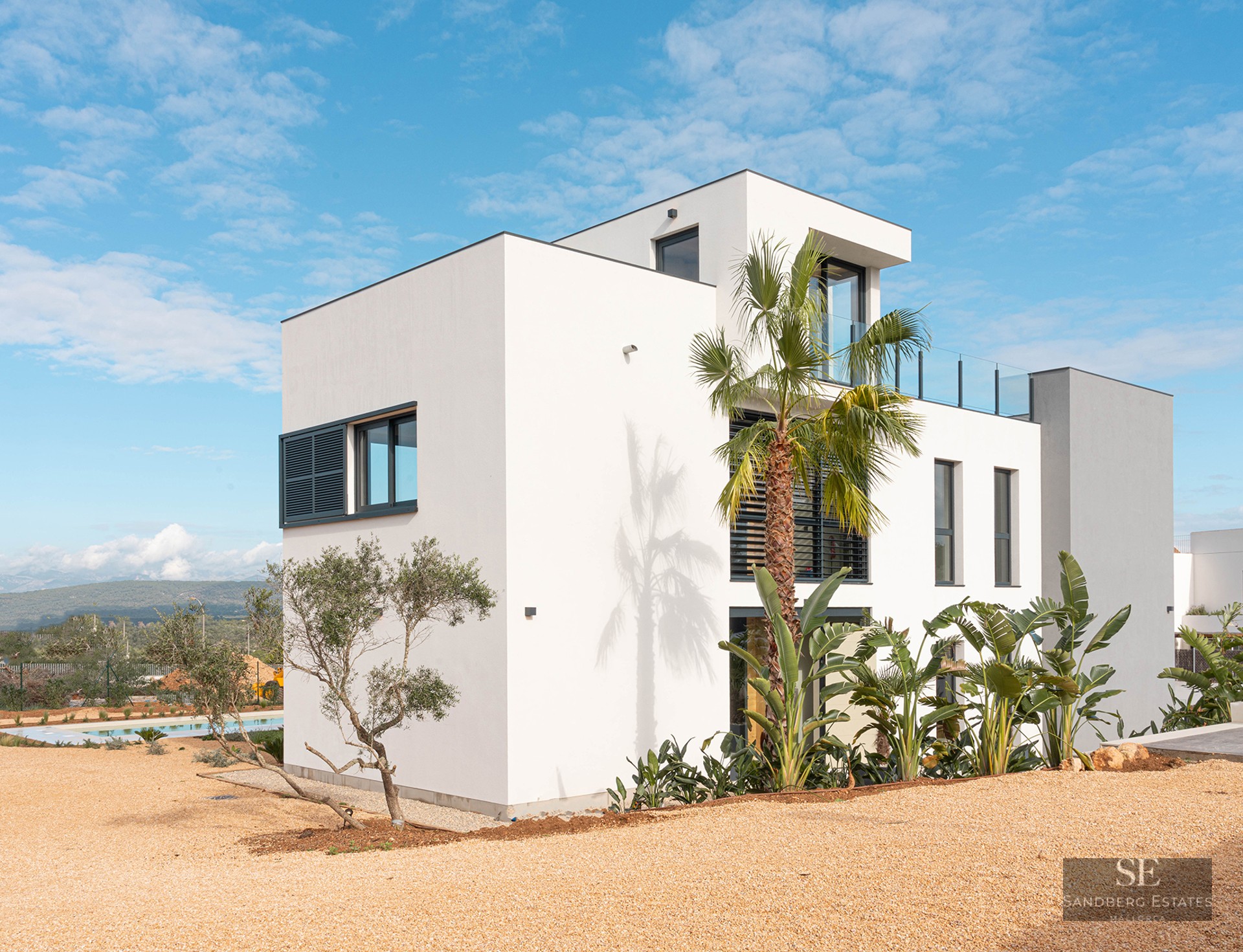 Exterior of a three-level white villa with modern architecture, palm trees, and a gravel yard under a blue sky.