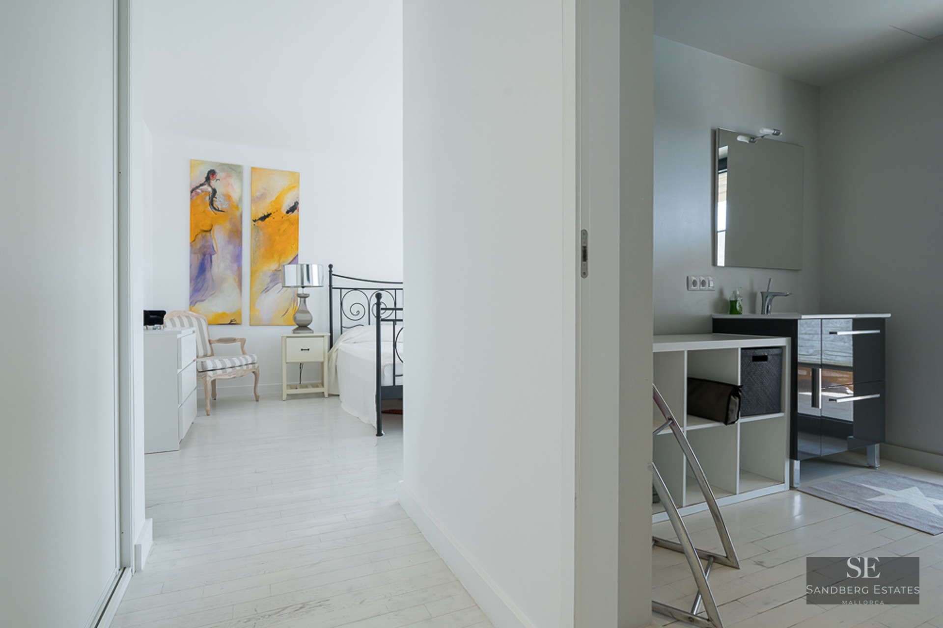 View from a bright hallway into a white bedroom with abstract art and an adjacent bathroom with a black vanity.