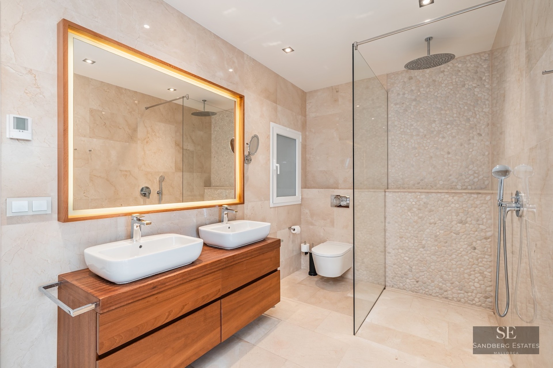 Modern master bathroom with double sinks on wood vanity, backlit mirror, and glass walk-in shower with pebble wall.