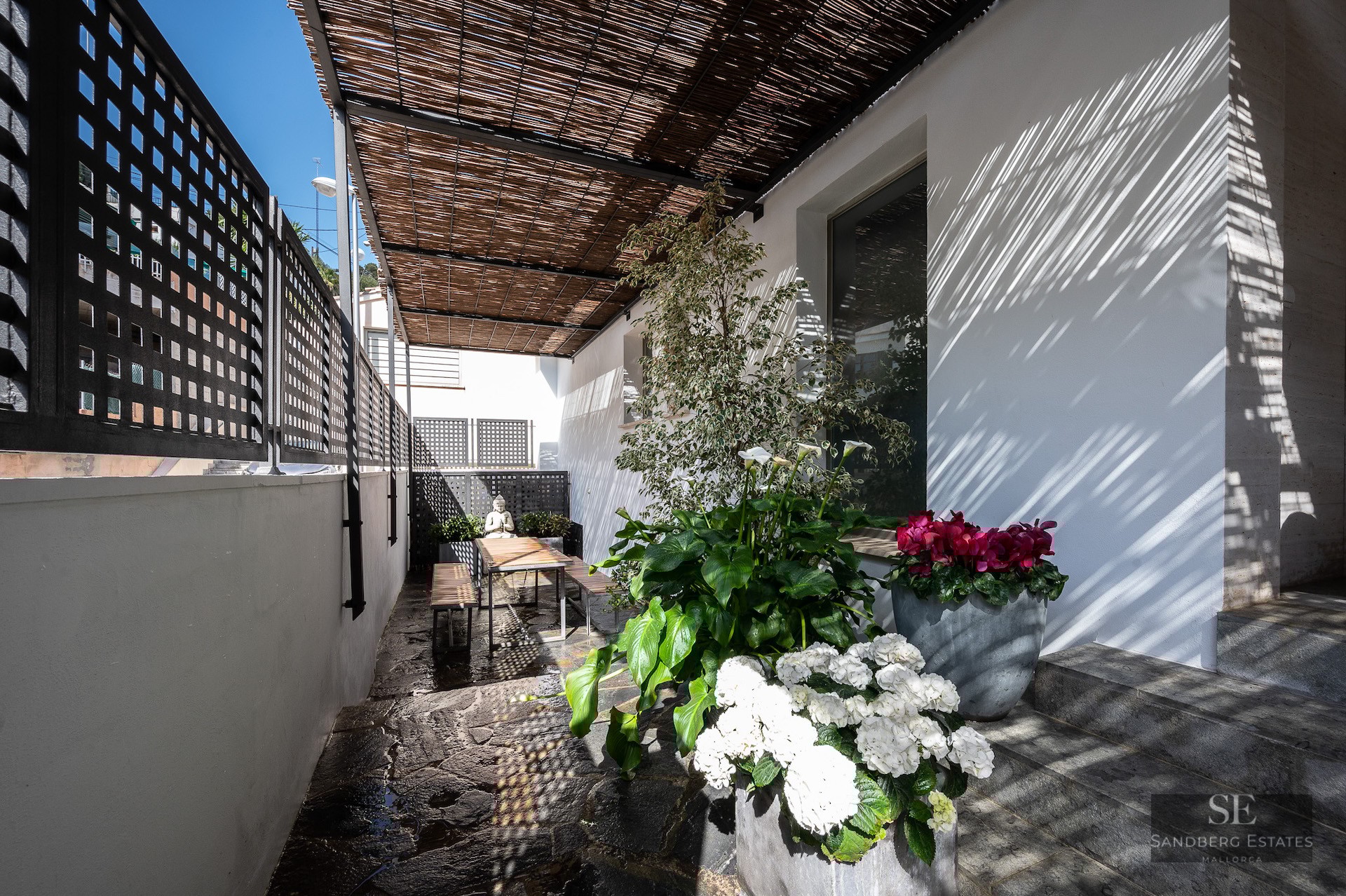Sunlit terrace with bamboo roof, stone floor, wooden dining set, and lush potted flowers against a white wall.