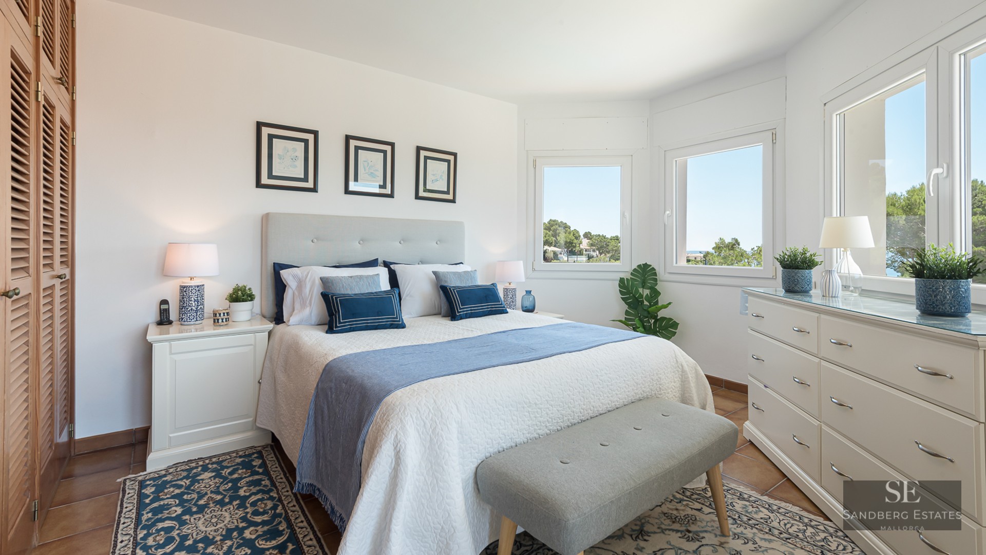 Bright bedroom with a white bed, blue pillows, white dresser, large windows, and terracotta tile floors.