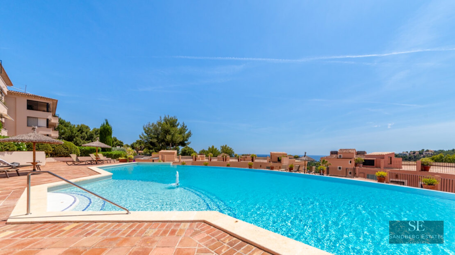 Grande piscine bleue incurvée avec fontaine, terrasse en terre cuite, parasols en paille et vue sur mer.