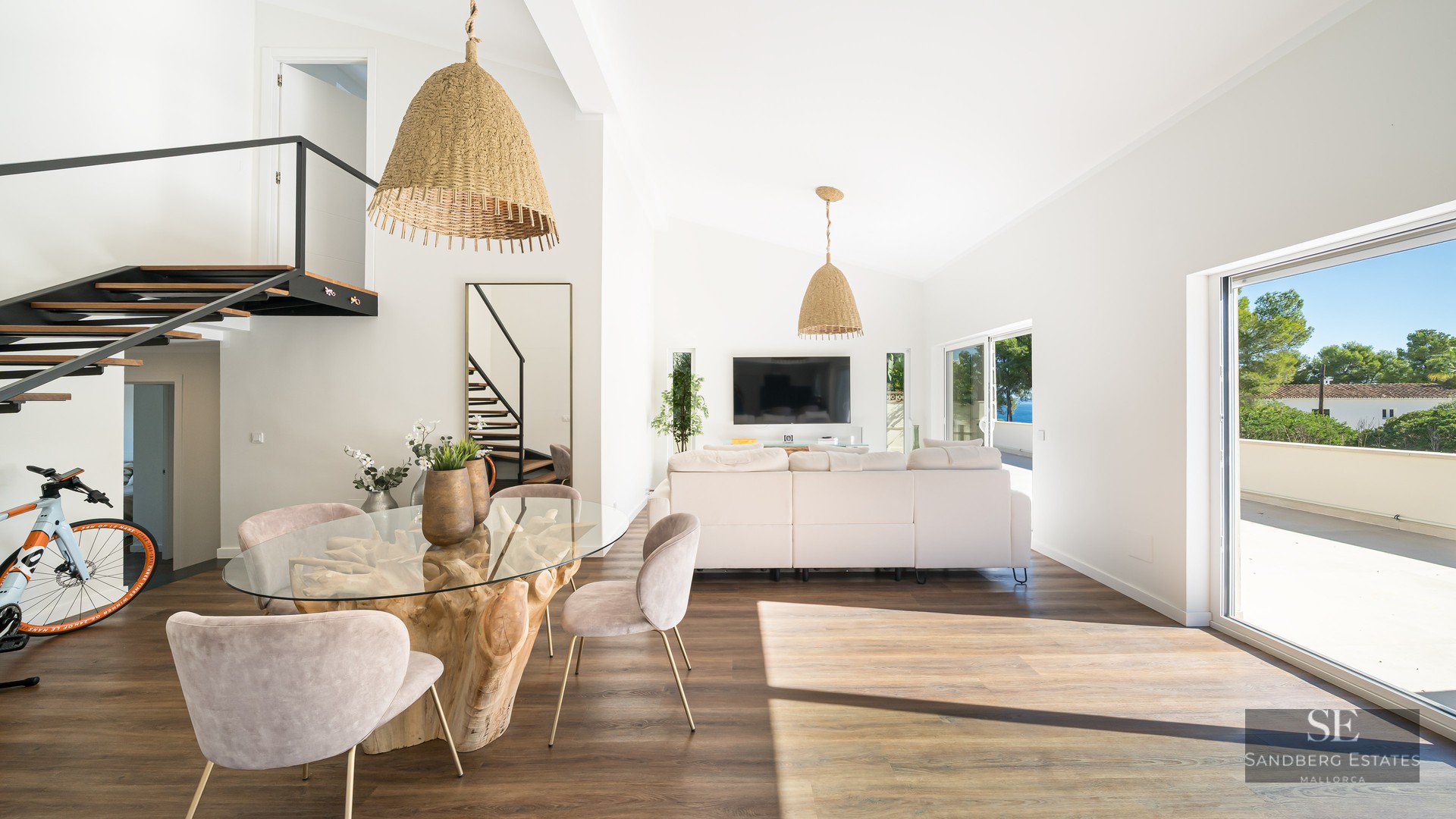 Salon lumineux avec table à manger en bois flotté, canapé blanc et suspensions en osier près de grandes baies vitrées.