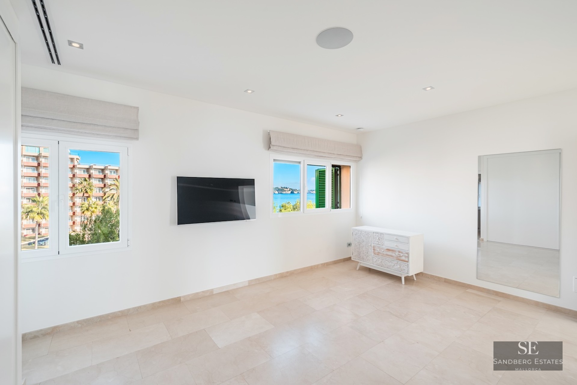 Bright room with white walls, stone floors, wall-mounted TV, and windows overlooking the Mediterranean sea.
