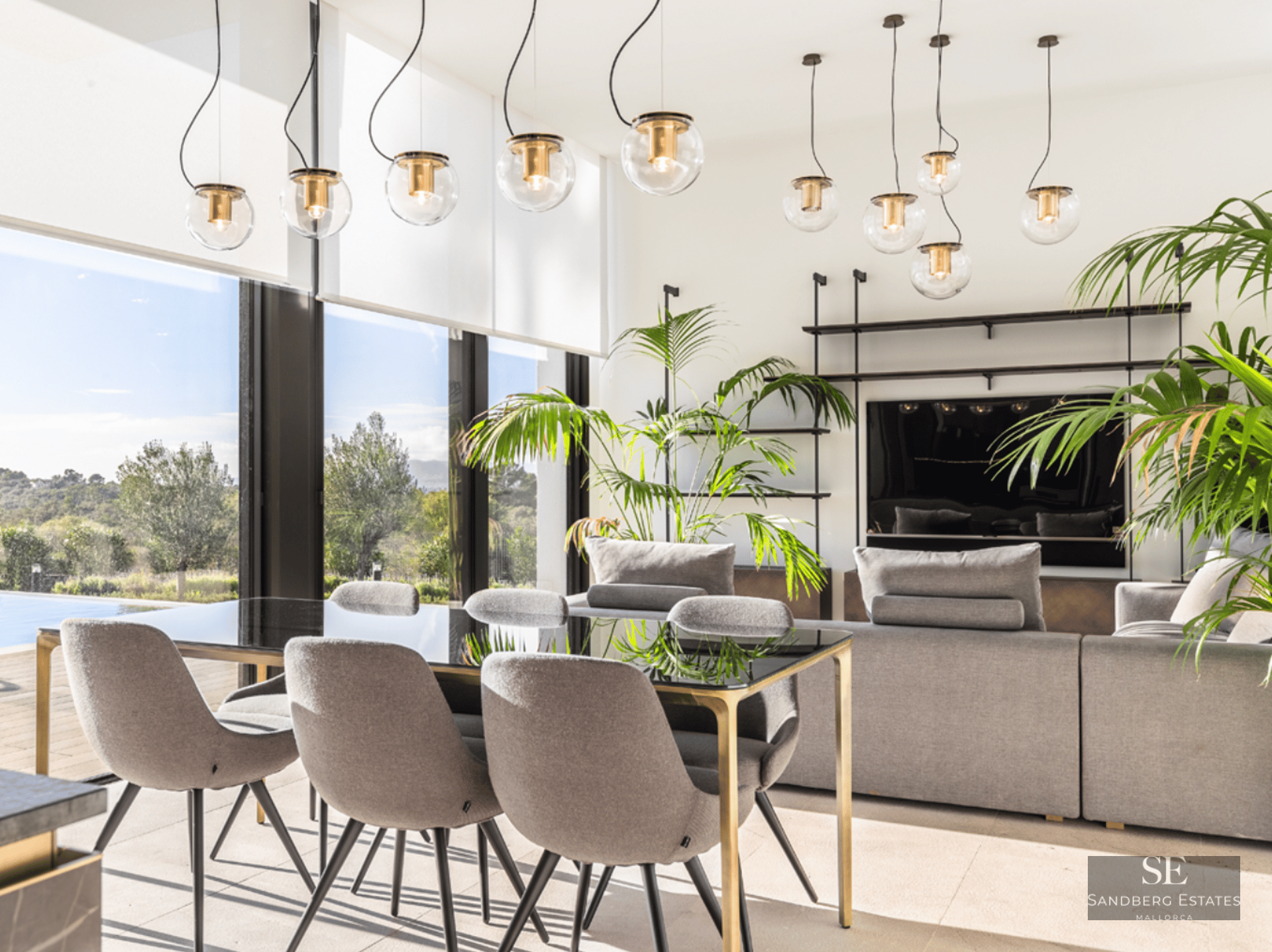 A luxurious modern dining area with a glass table and grey chairs, overlooking a living room and outdoor greenery.