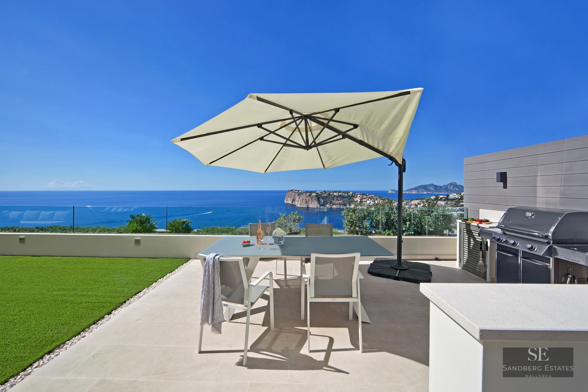 Outdoor terrace with dining table, sun umbrella, BBQ grill, and stunning ocean views under a clear blue sky.