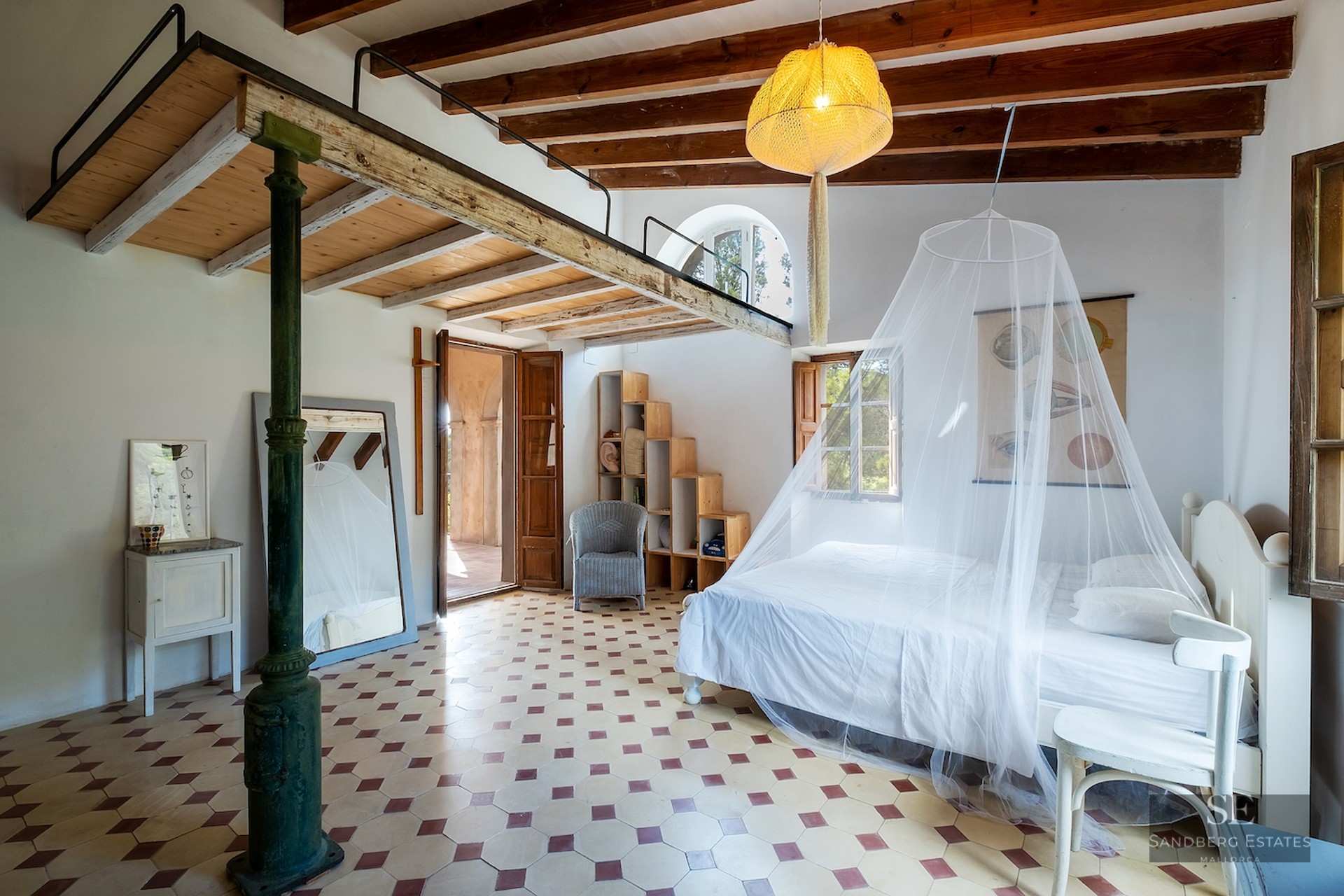 Spacious bedroom with white bed, mosquito net, wooden mezzanine loft, exposed beams, and decorative tiled floor.
