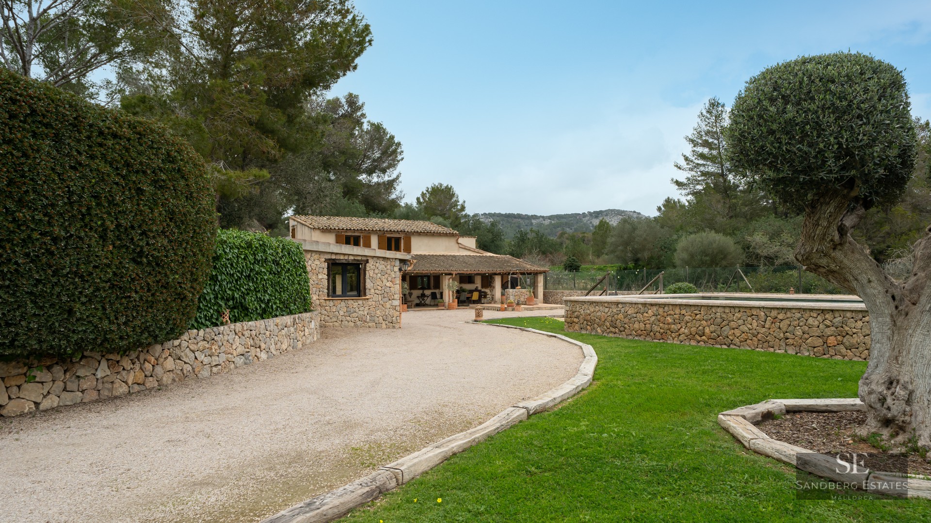A rustic stone house with a gravel driveway, lush green lawn, and an elevated stone pool area under a clear sky.
