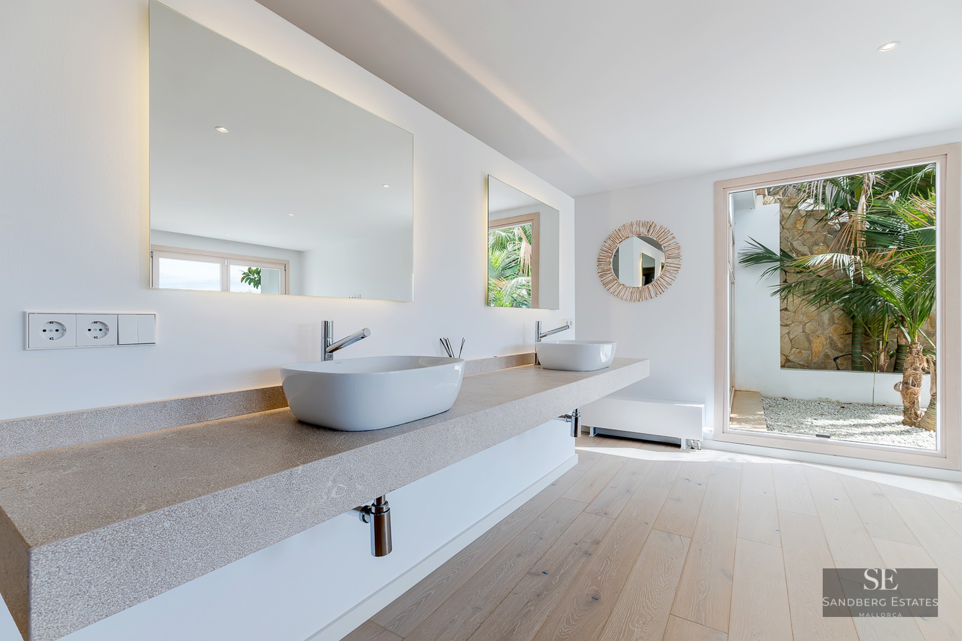 Bright bathroom with stone floating vanity, two white sinks, large mirrors, and a window to a tropical garden.