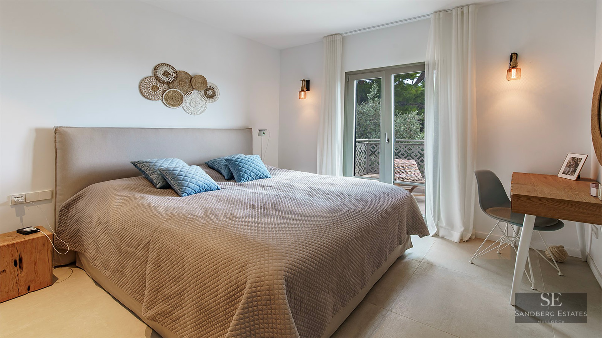 Modern bedroom featuring a large bed, beige textured bedding, blue pillows, wooden desk, and glass door to a balcony.