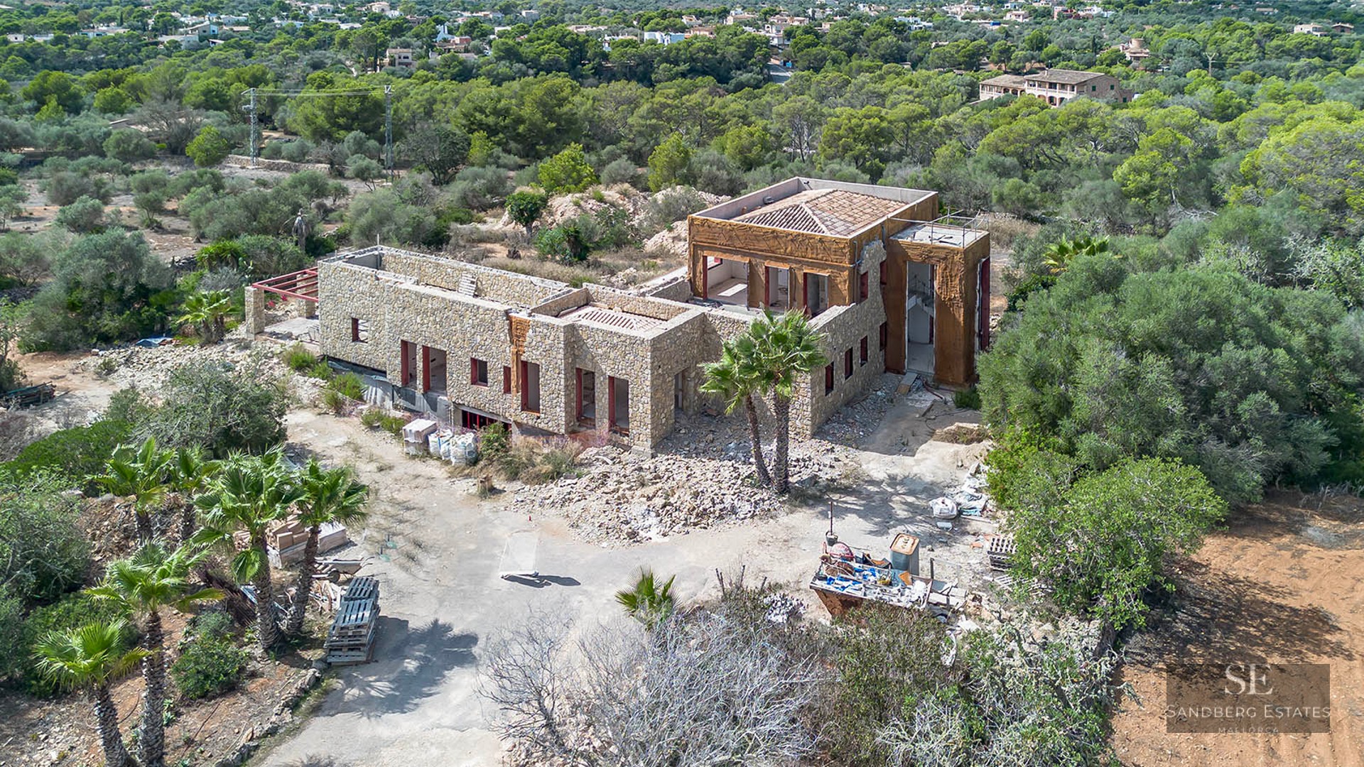 Elevated view of a large stone villa under construction surrounded by dense Mediterranean greenery.