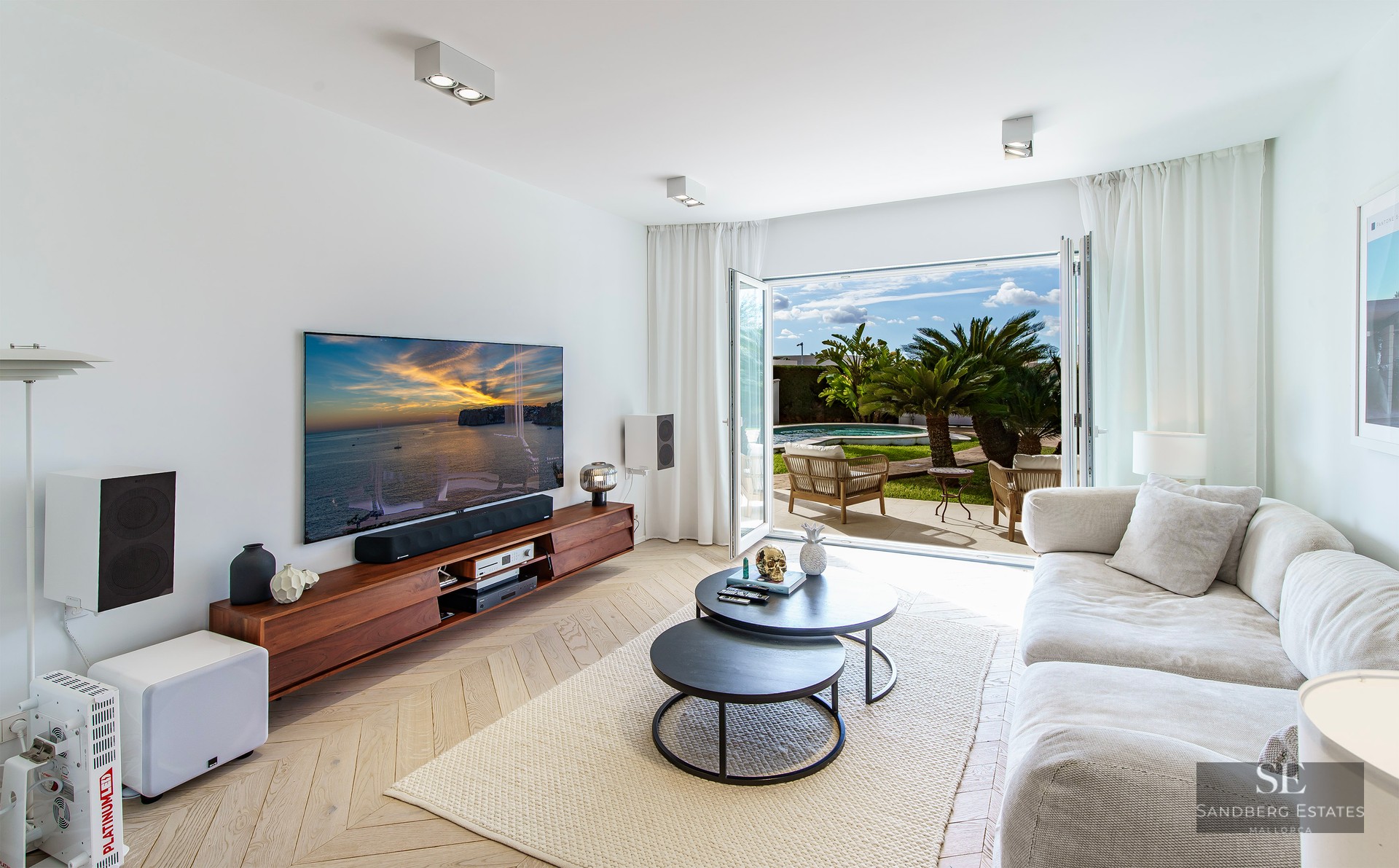 Bright living room with white sofa, herringbone wood floor, and open doors to a sunny terrace with palms.