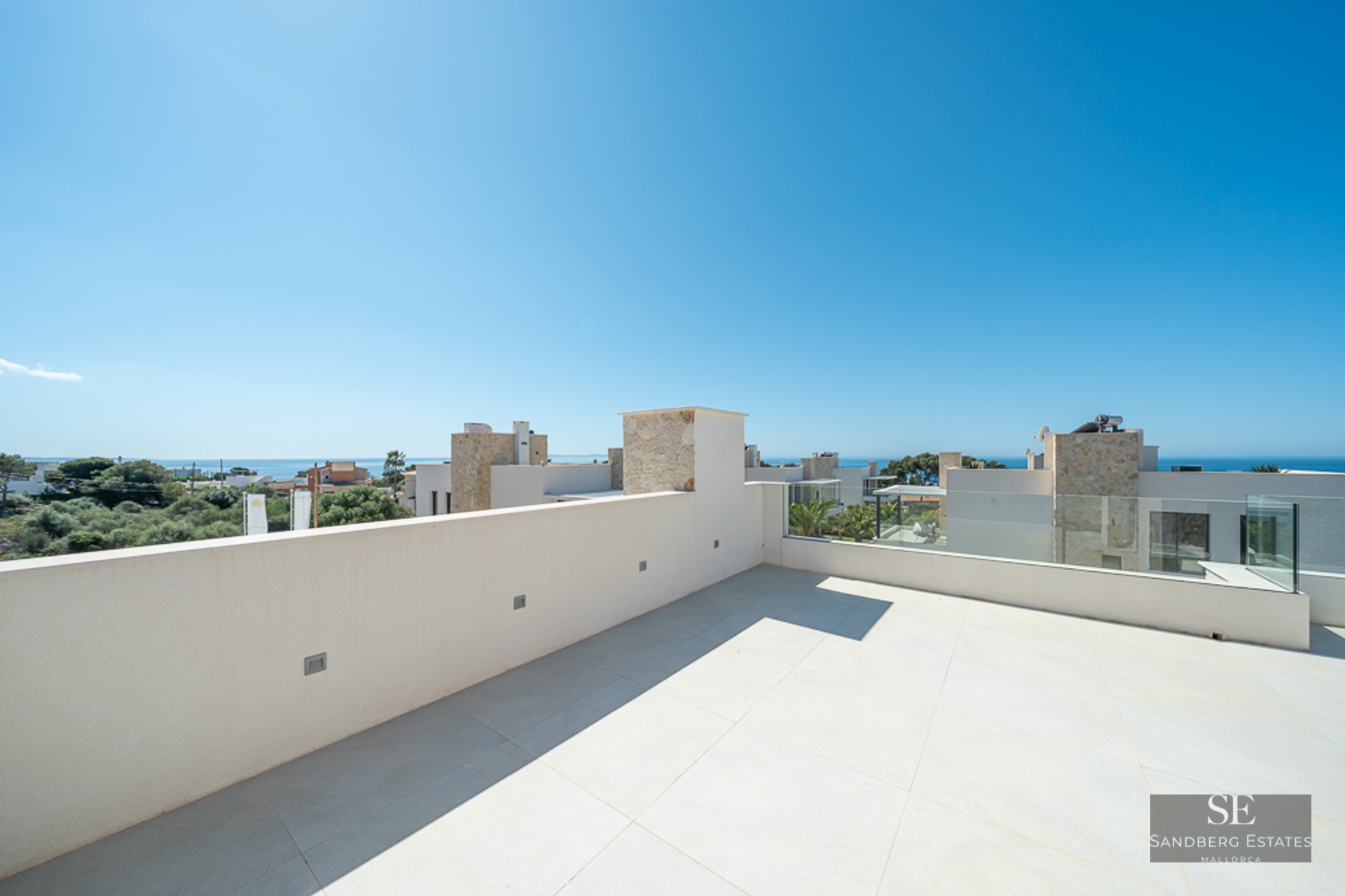 Wide white-tiled rooftop terrace with a low wall and glass railing, overlooking modern villas and the ocean.