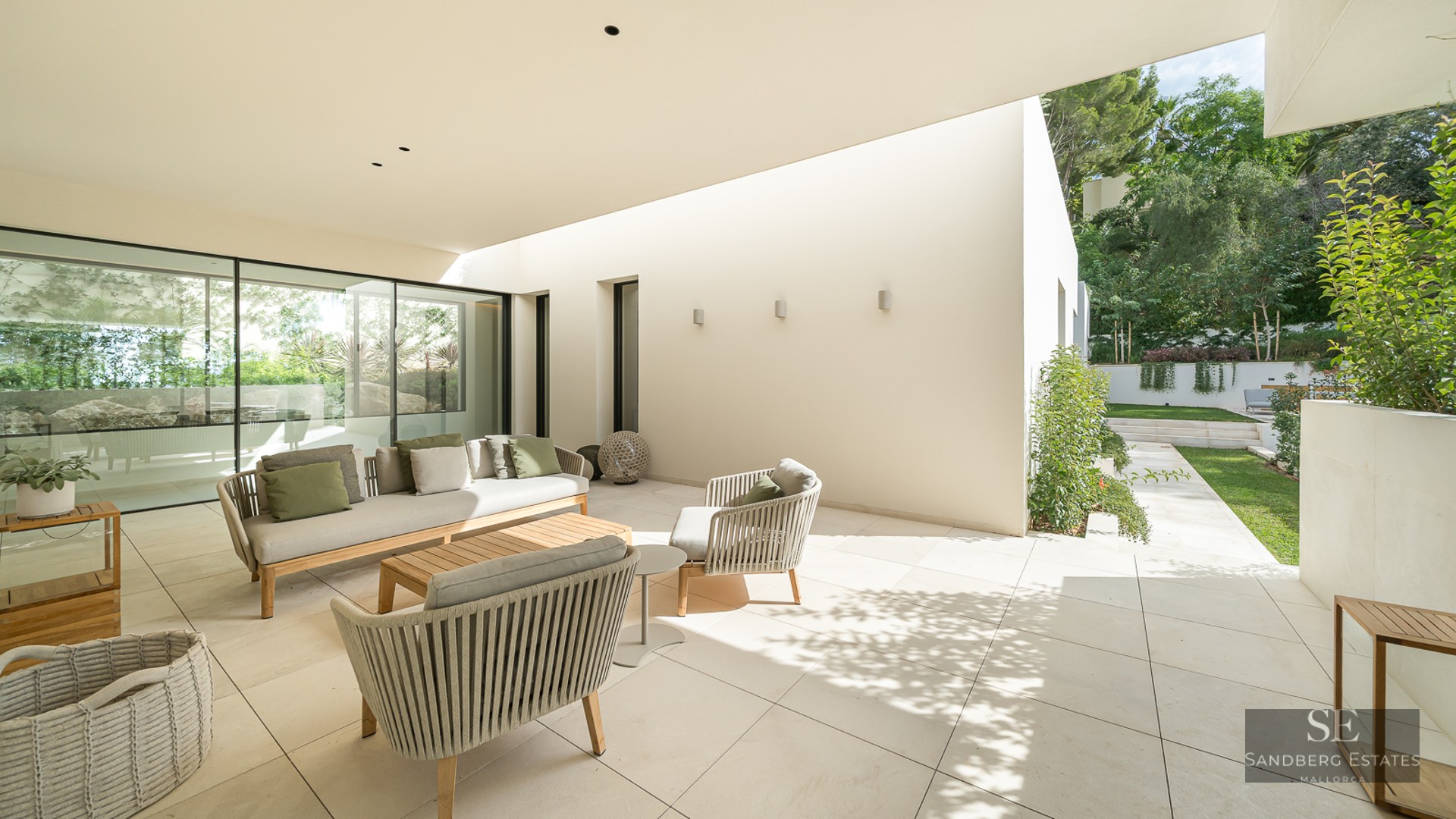 Luxury outdoor terrace with a beige sofa, green cushions, and woven chairs on a stone patio overlooking a garden.