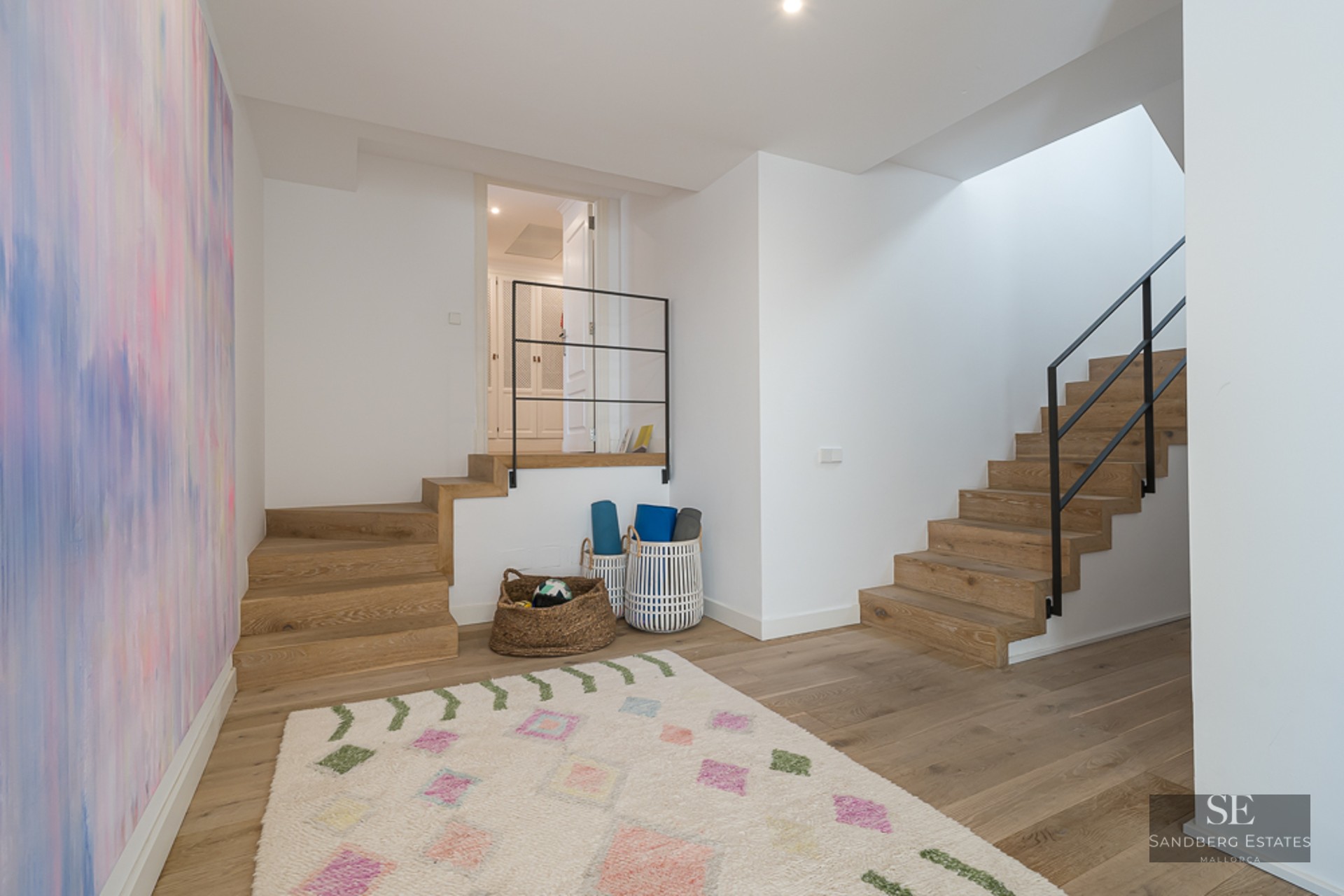 Pièce blanche avec deux escaliers en bois, fresque colorée et tapis de yoga dans des paniers.