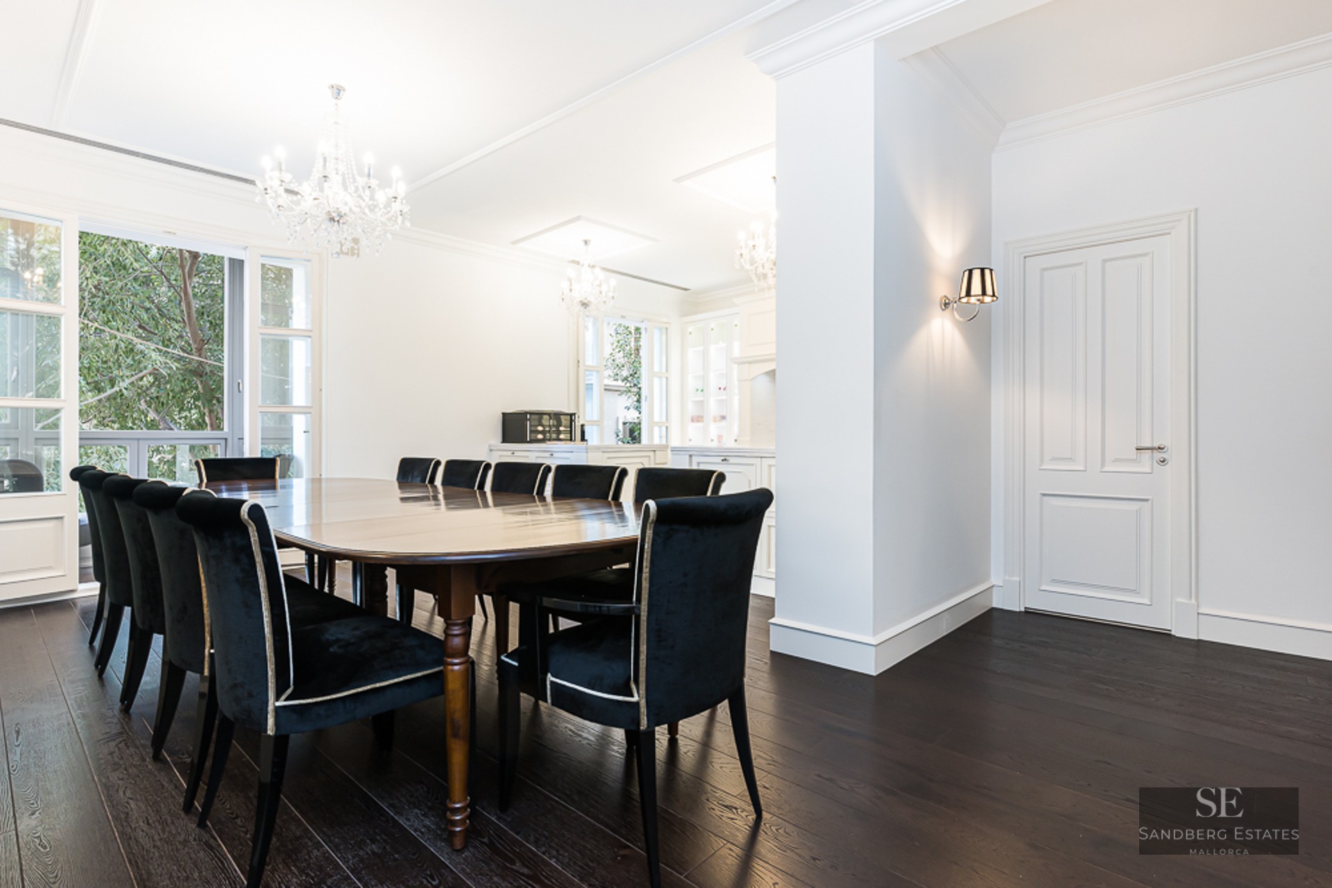 A large dark wood dining table with black velvet chairs under crystal chandeliers in a bright white room.