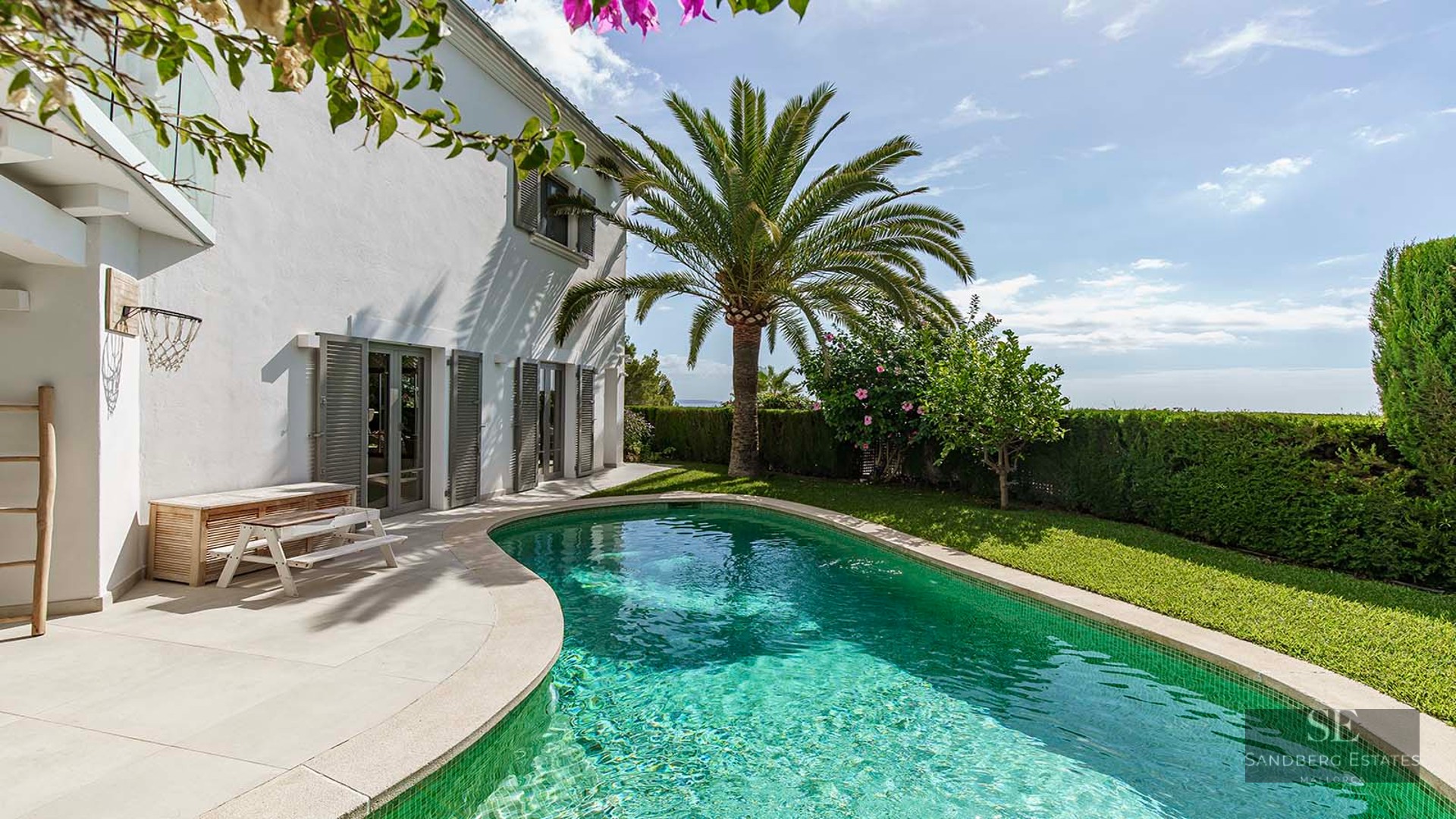 A curved turquoise pool bordered by a lush lawn, a palm tree, and a white villa with grey shutters under a blue sky.