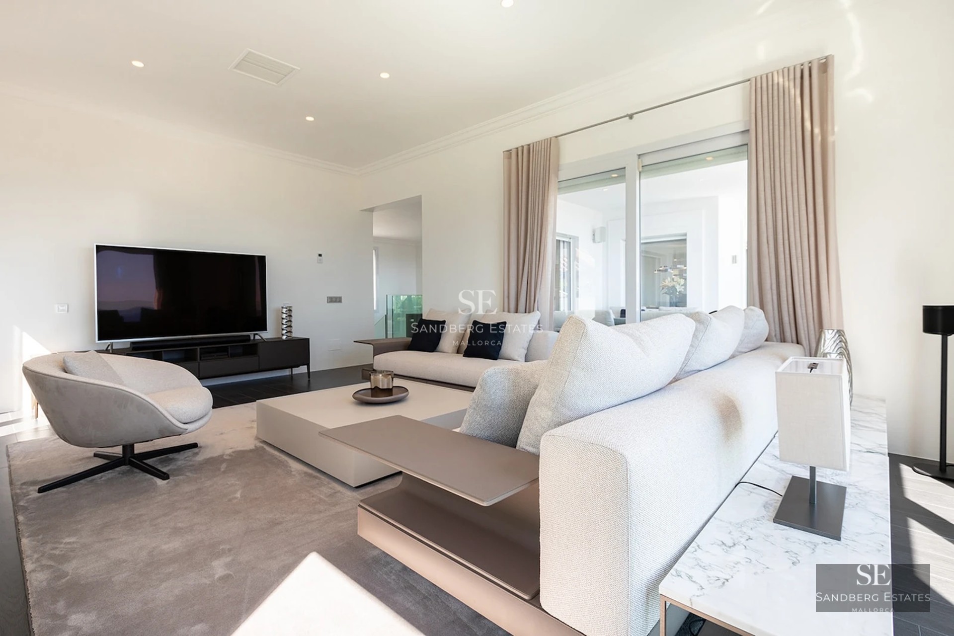 Modern living room with large grey sofas, coffee table, big screen TV, and floor-to-ceiling glass doors.