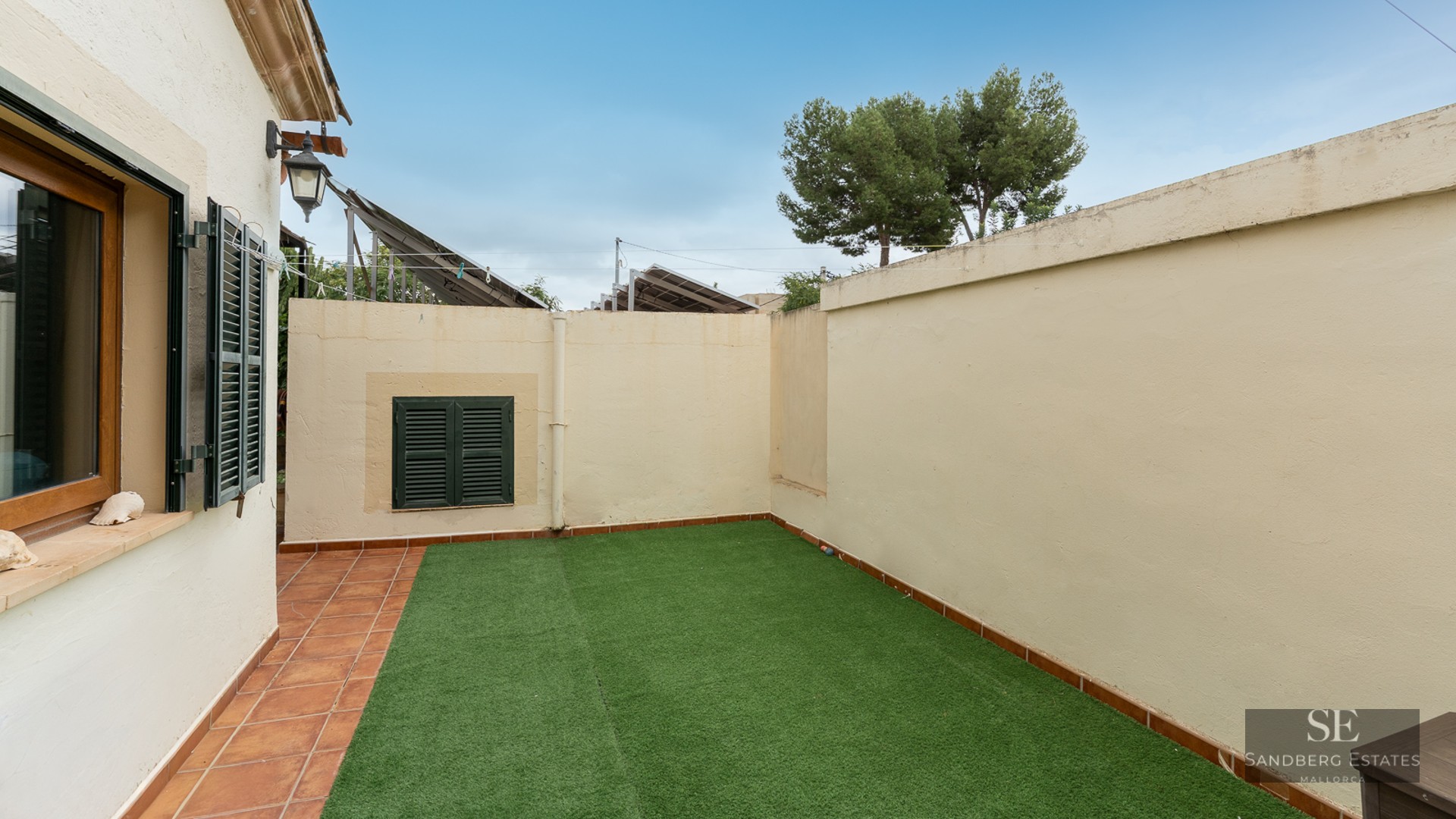 Small walled garden with artificial grass and terracotta tile border under a blue sky.