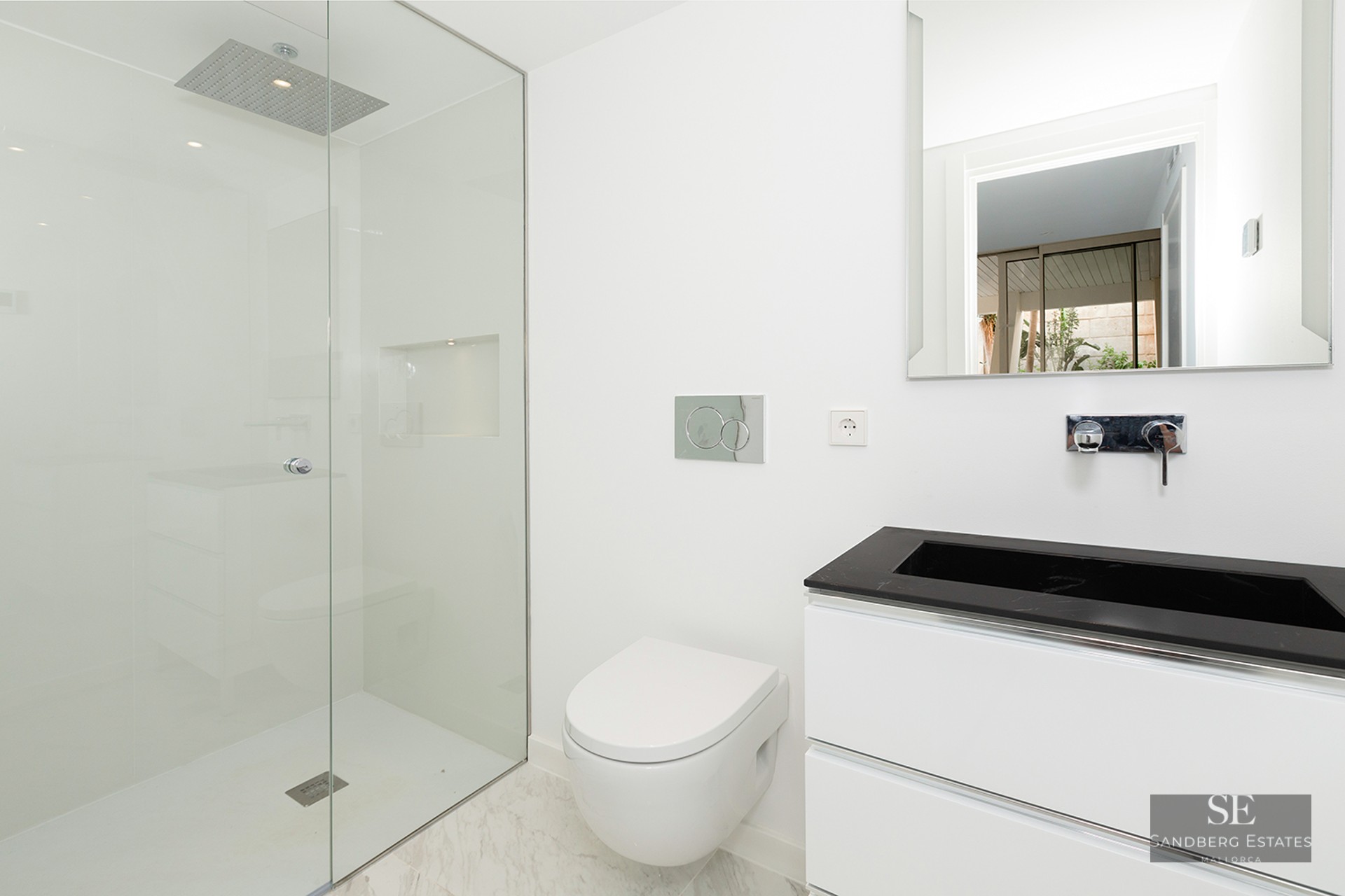 A bright, minimalist bathroom featuring a glass walk-in shower, a white wall-hung toilet, and a black and white vanity.