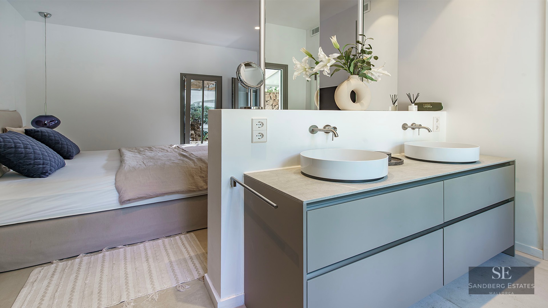 Open-plan master bedroom featuring a double vanity with two vessel sinks and a large bed with blue pillows.