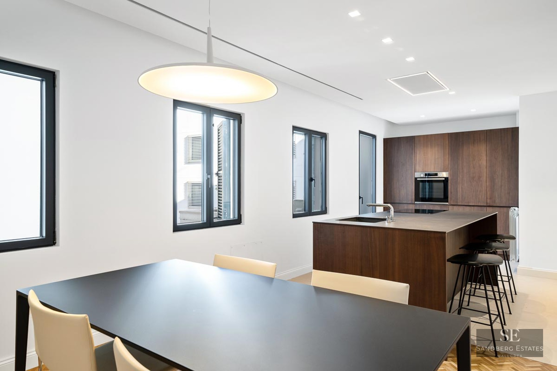 Open-plan kitchen featuring a dark wood island, black dining table, white walls, and three rectangular windows.