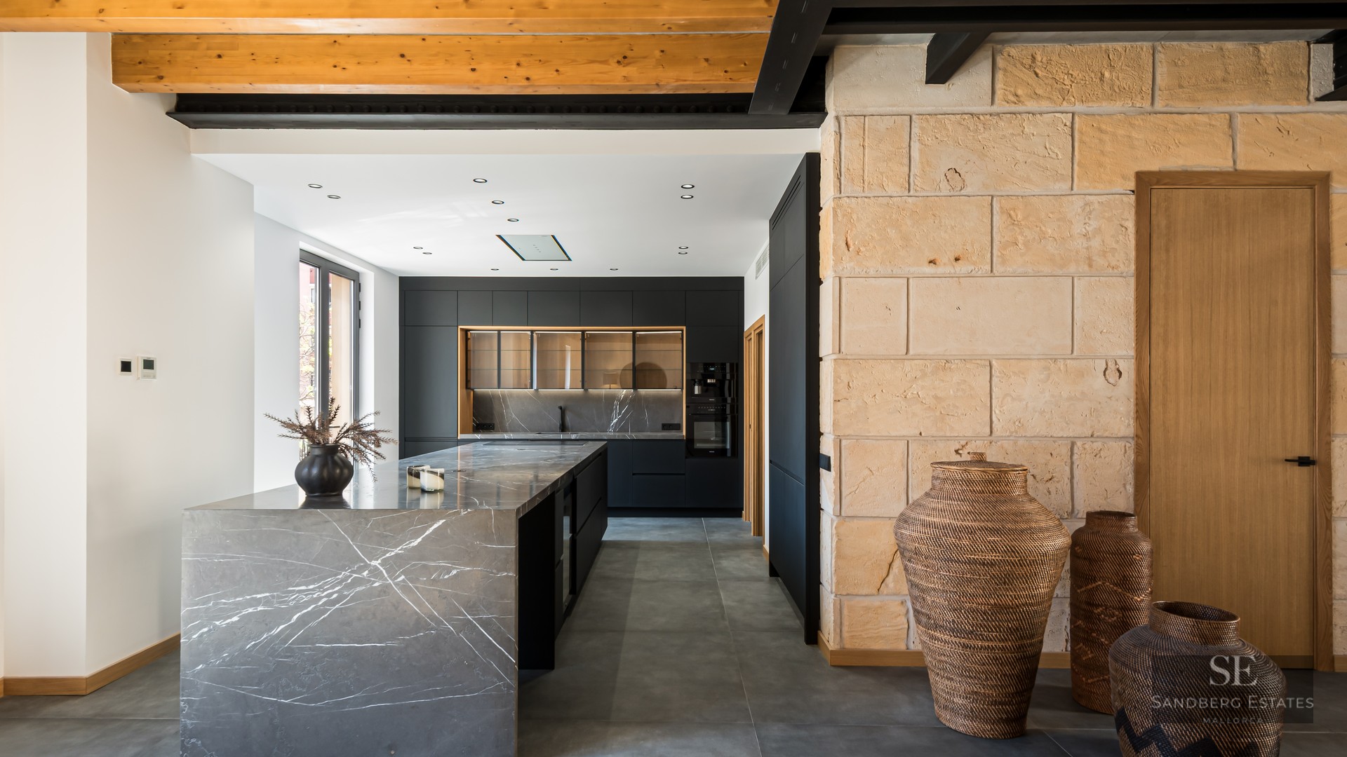 A contemporary kitchen featuring a grey marble island, black cabinetry, exposed wooden ceiling beams, and a stone wall.