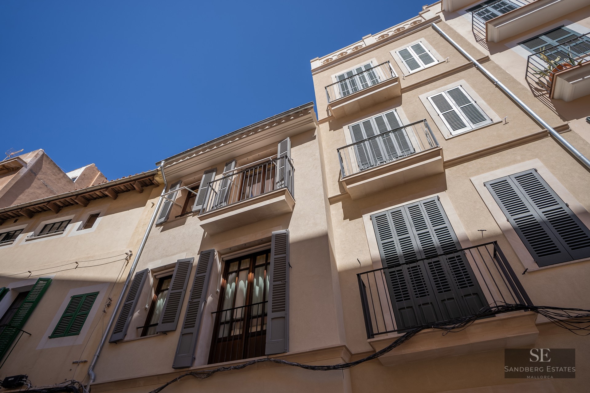 Low angle view of a beige building facade with green shutters and wrought iron balconies against a clear blue sky.