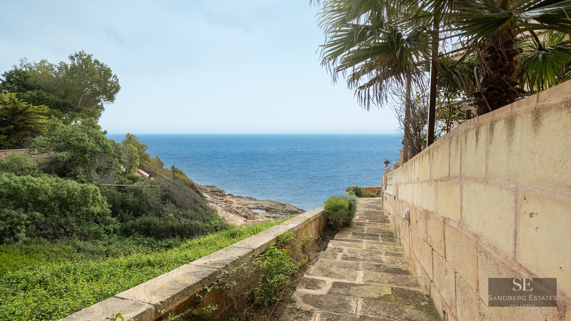 A stone pathway between a wall and greenery leads directly towards a vast, blue ocean.