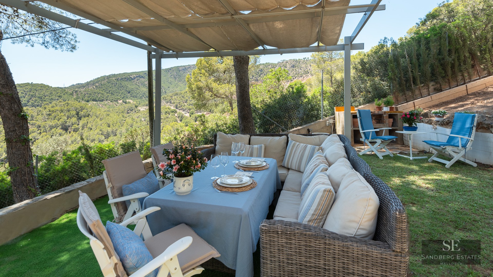 Outdoor dining area under a pergola with wicker sofas and views of green forested hills.