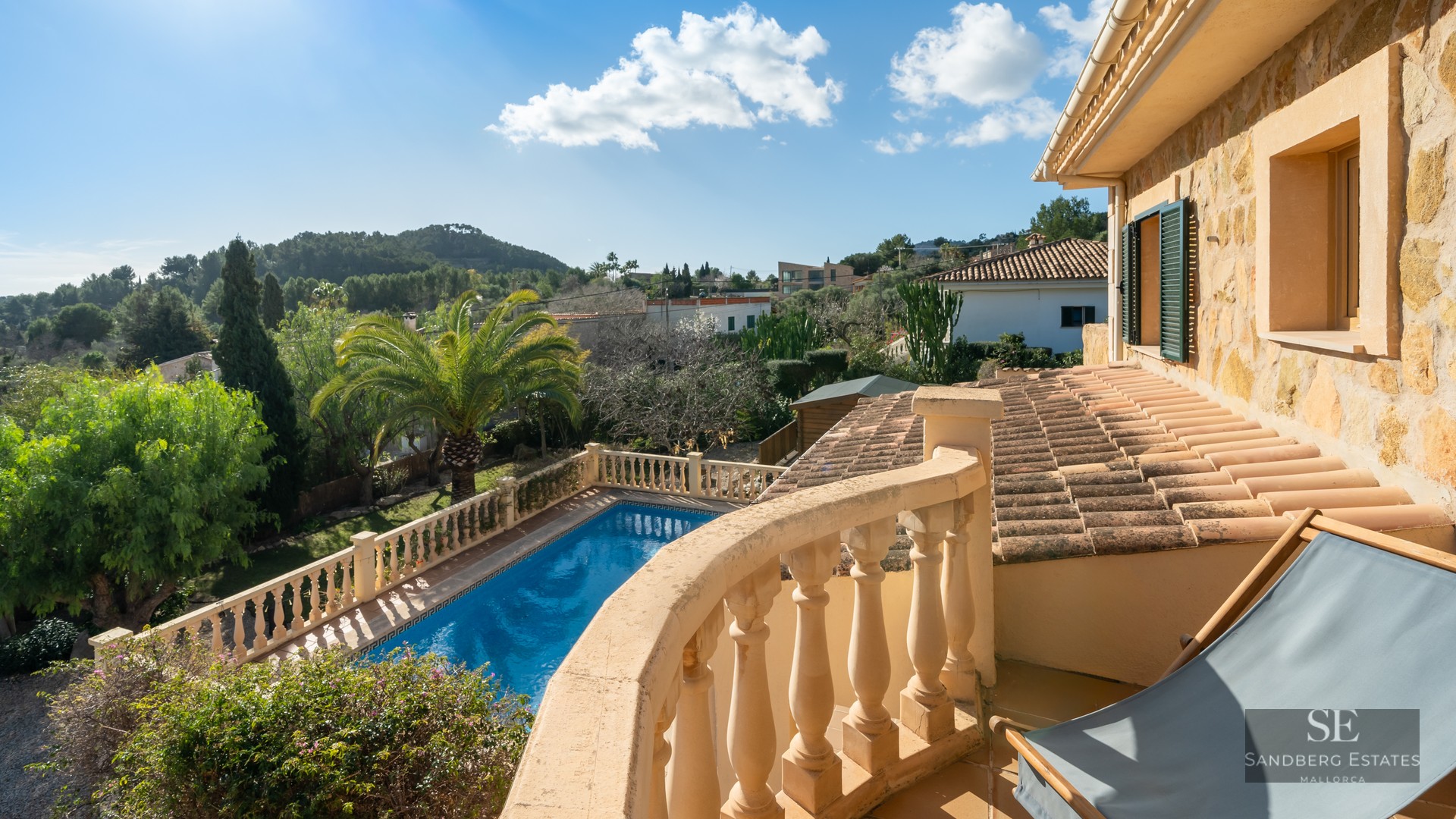 View from a stone balcony overlooking a blue swimming pool, lush gardens, and rolling hills under a clear blue sky.