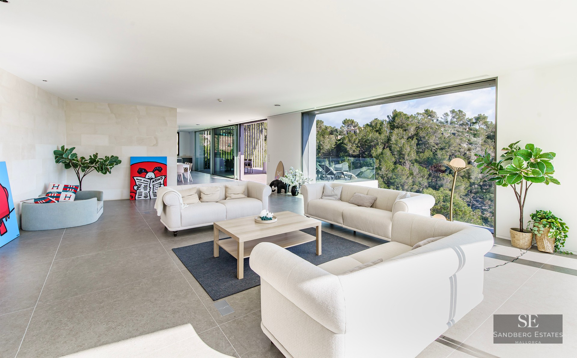 Spacious modern living room with cream sofas and large glass walls overlooking a lush green forest.
