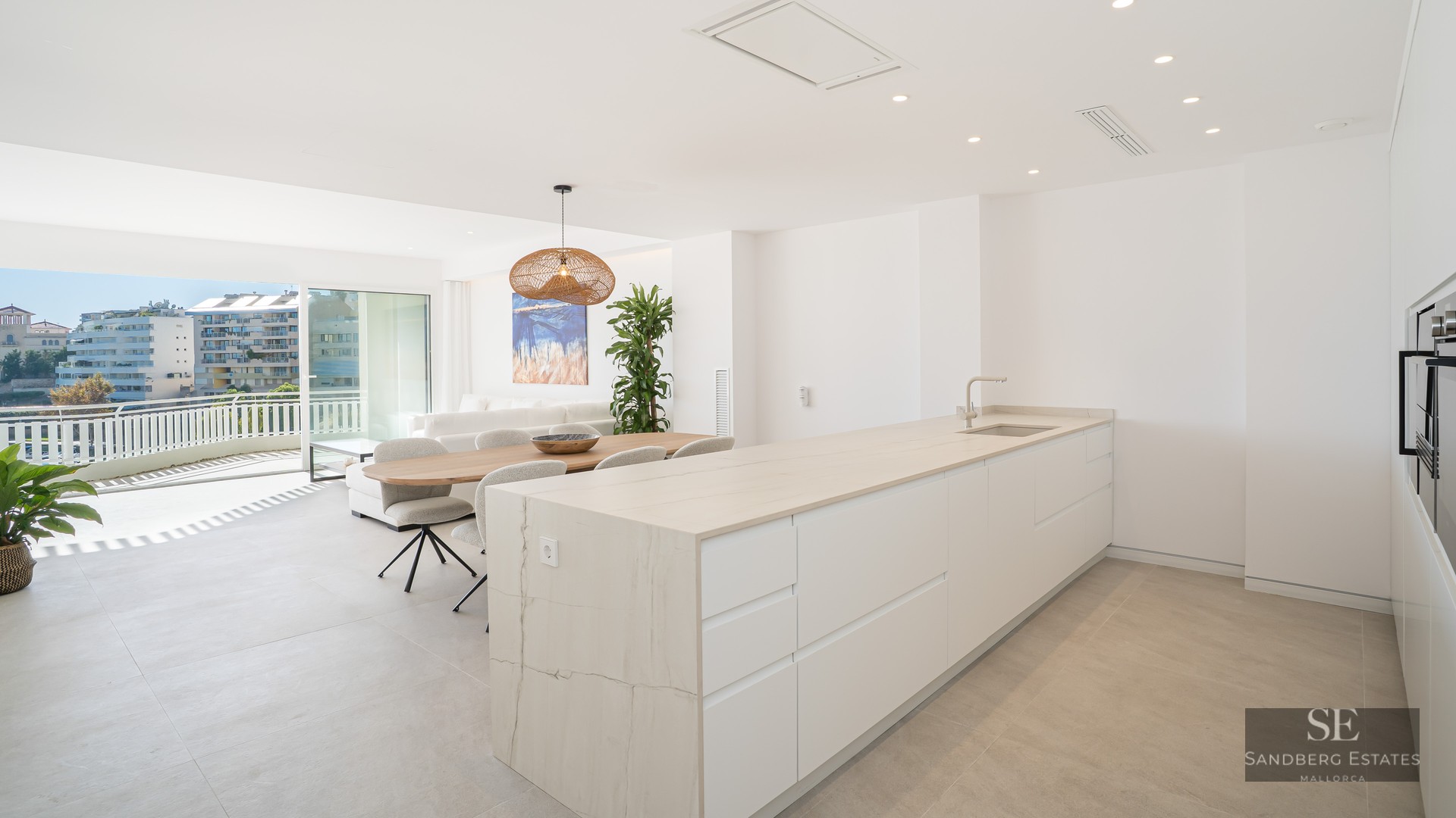 Modern white kitchen island and wooden dining table in a bright open-plan apartment with terrace and city views.
