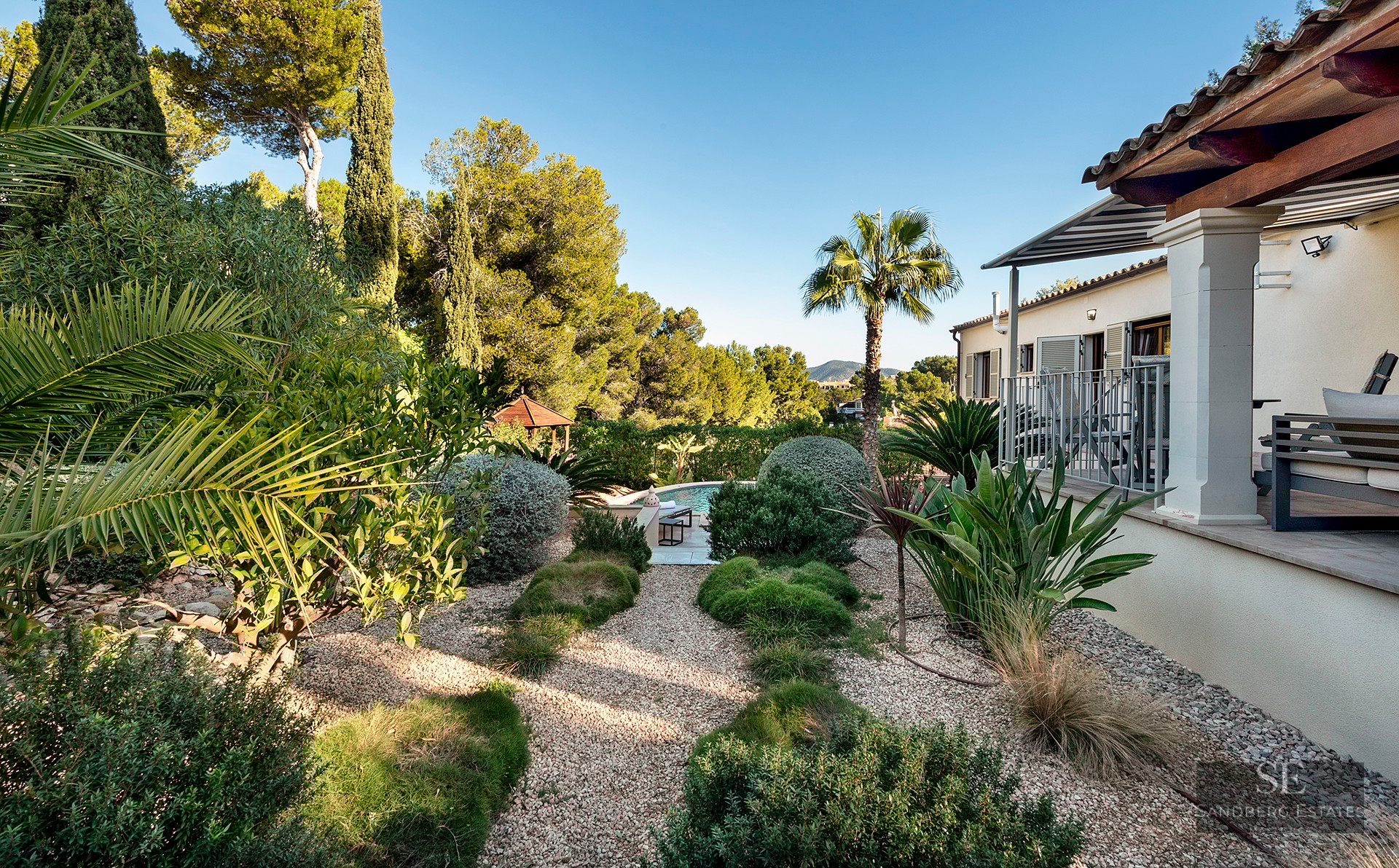 Lush landscaped garden with gravel paths leading to a swimming pool, next to a Mediterranean style villa.