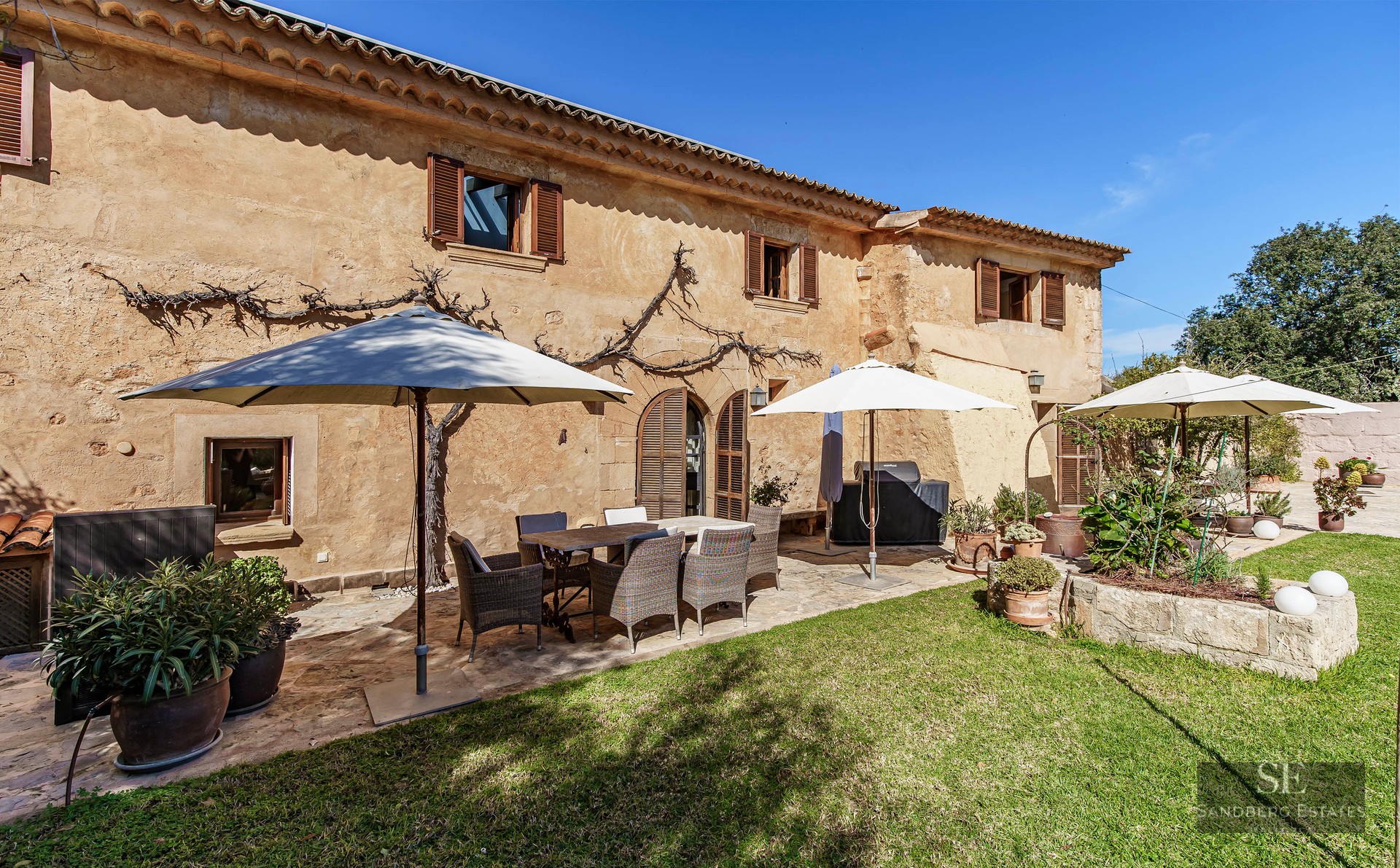 Sunny stone terrace with a dining table, wicker chairs, and white umbrellas next to a green lawn.