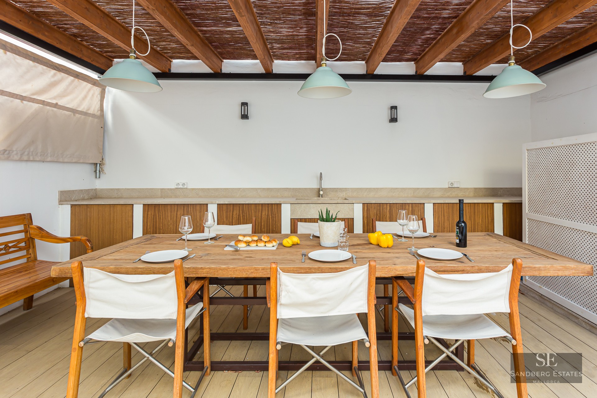 Covered terrace with large wooden table, white canvas chairs, and an outdoor kitchenette under a wooden pergola.