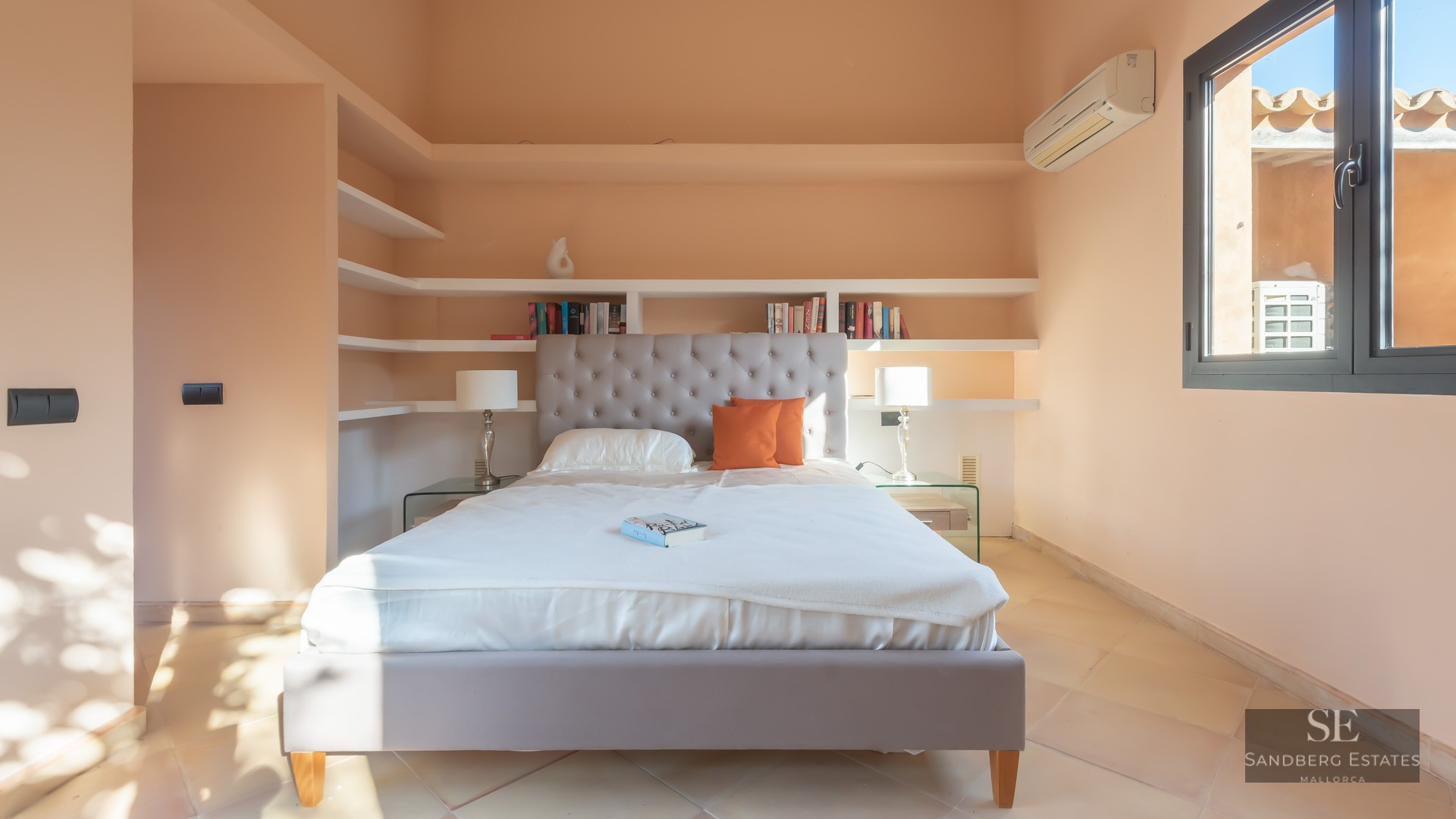 Bright bedroom featuring a tufted grey headboard, white bedding, built-in shelving, and terracotta tile floors.