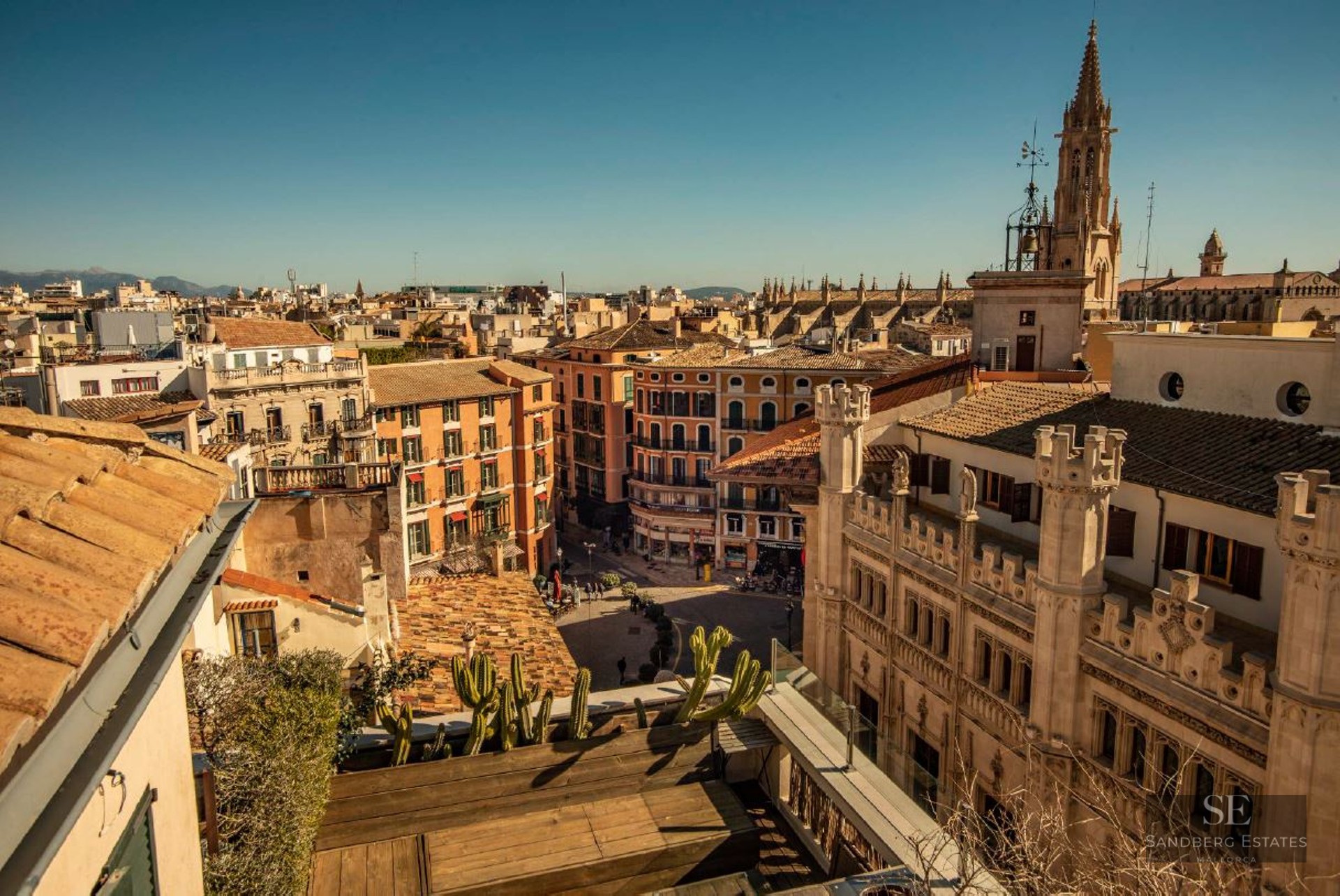 High-angle view of Mediterranean city rooftops and a Gothic cathedral spire under a bright blue sky.
