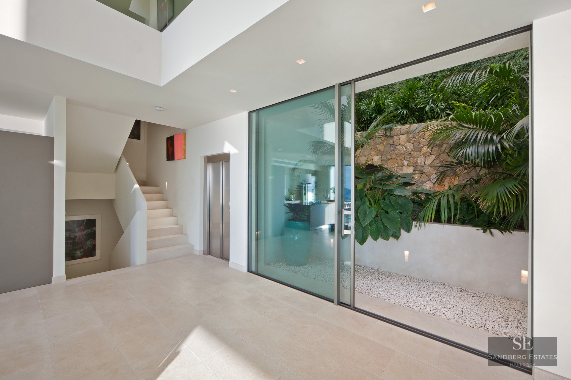 Minimalist lobby with light stone floors, an elevator, white stairs, and a glass door to a lush tropical patio.