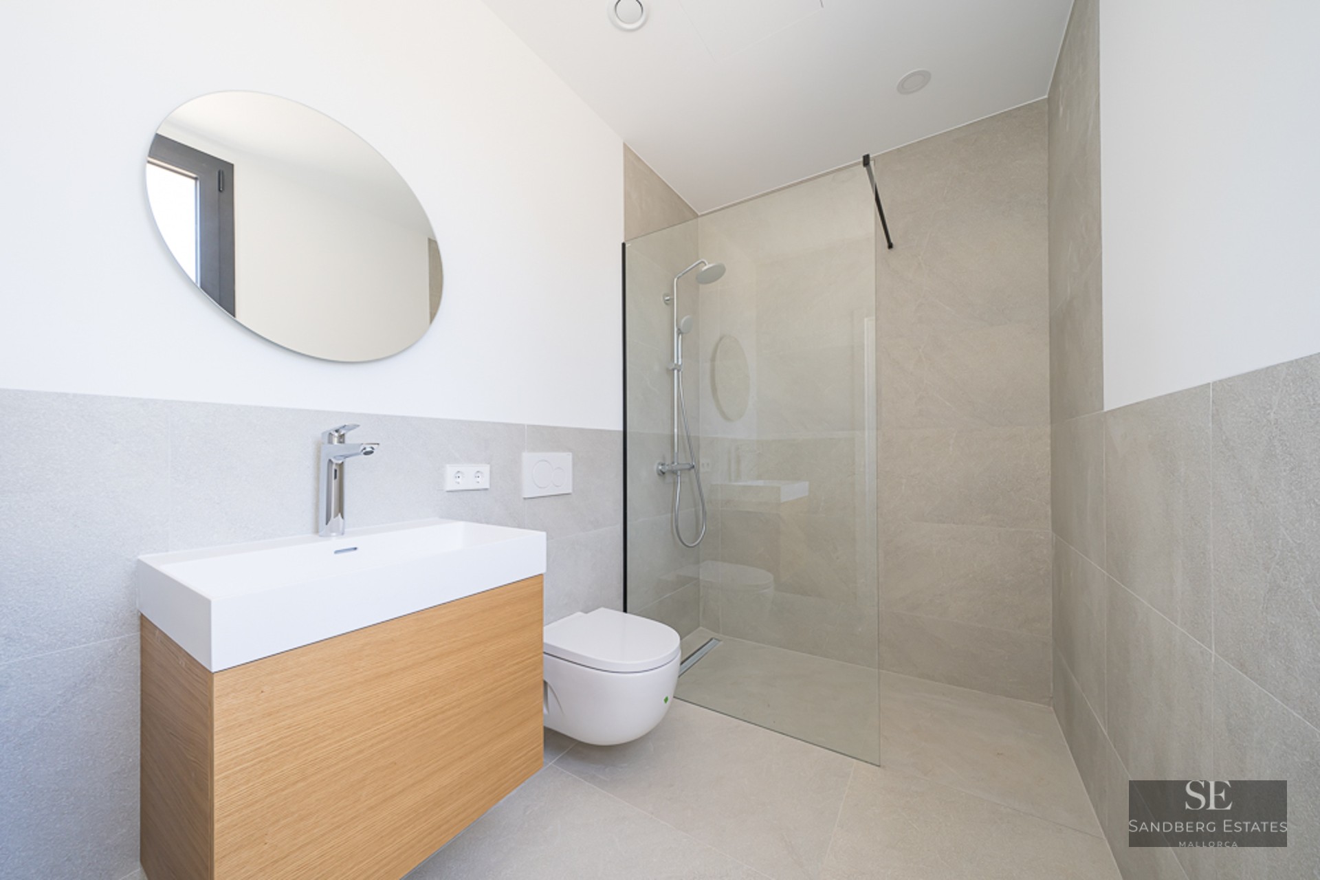Minimalist bathroom with light wood vanity, round mirror, white toilet, and glass walk-in shower with beige tiles.