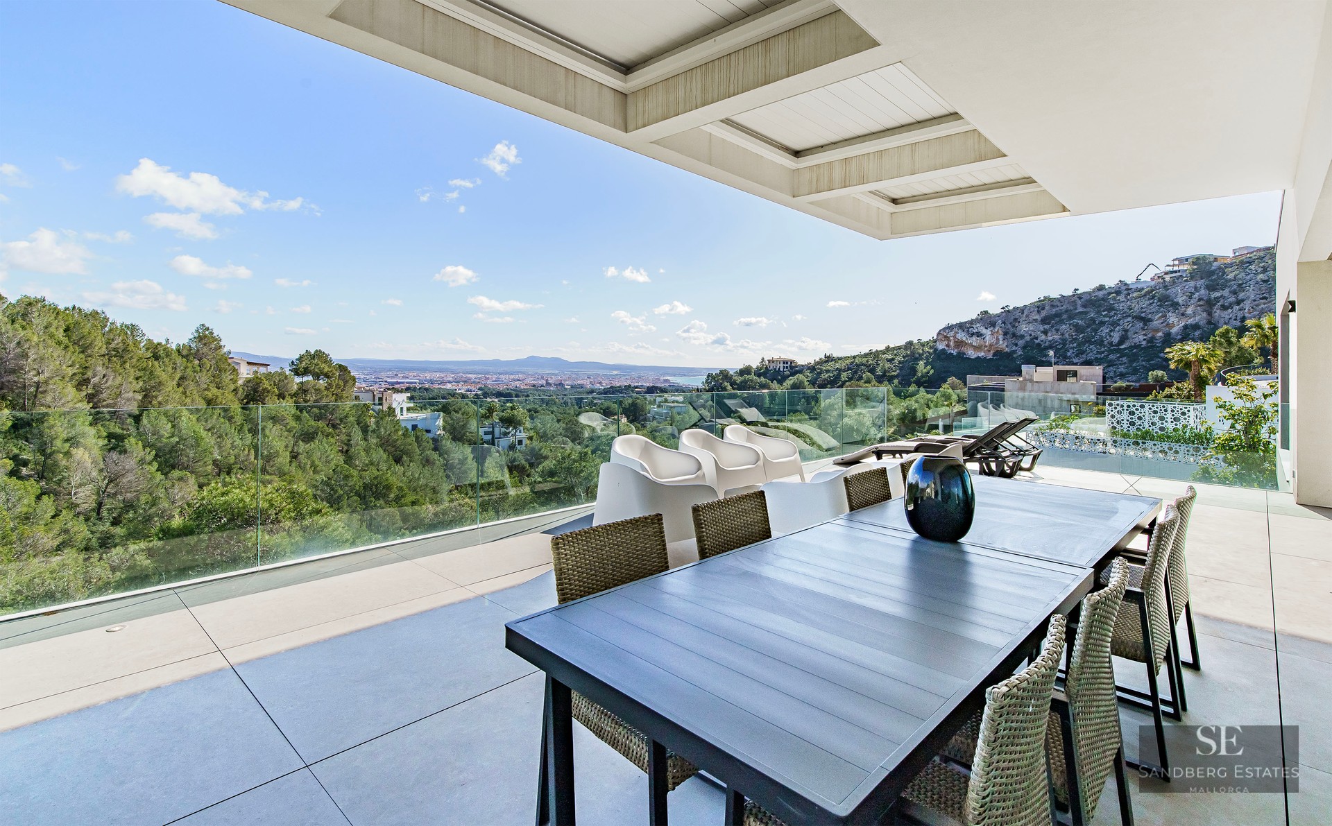 Modern terrace with a dining table and glass railings overlooking a lush green forest and distant city.