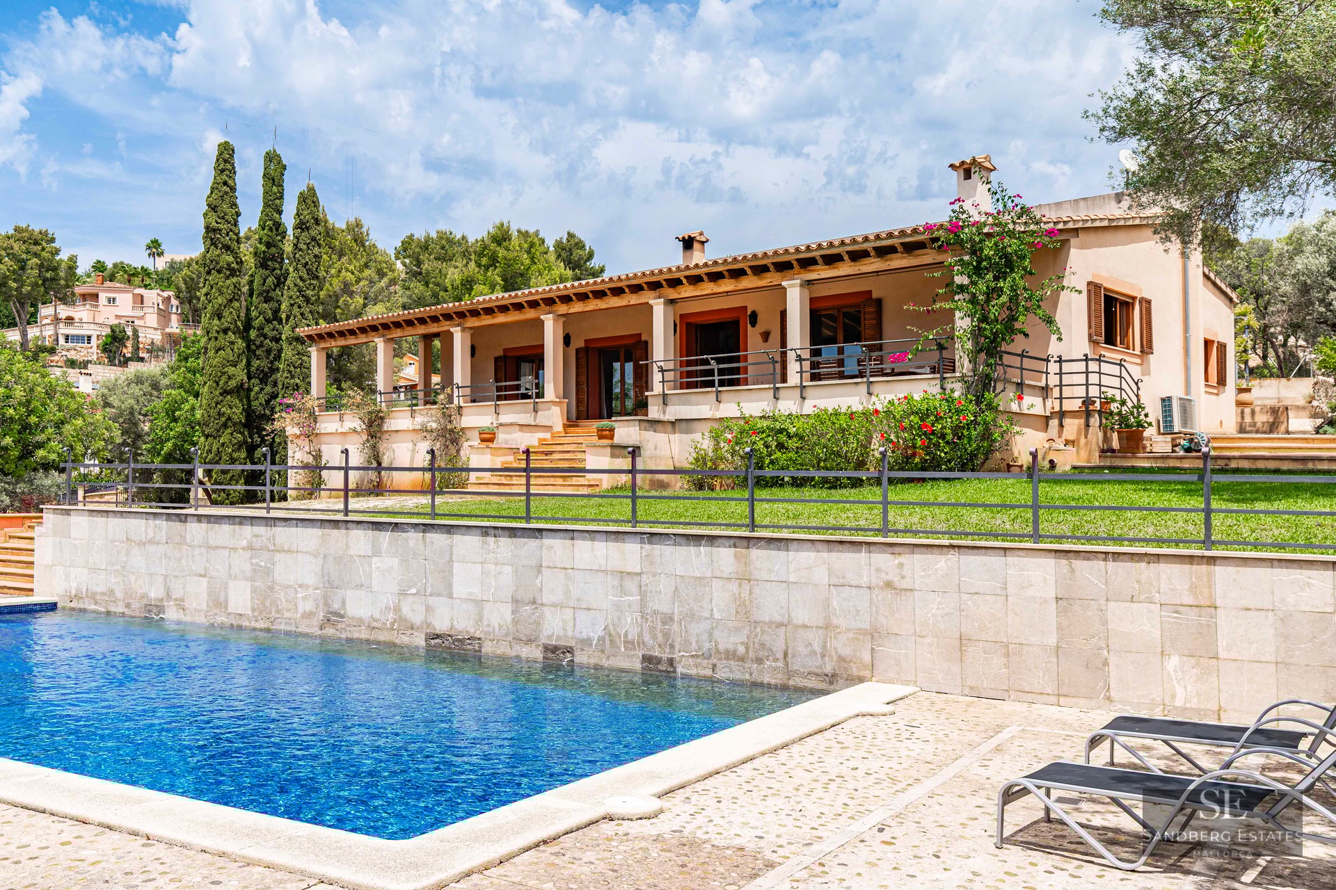 Large blue swimming pool and sun loungers in front of a Mediterranean villa with a stone wall, lawn, and covered terrace.